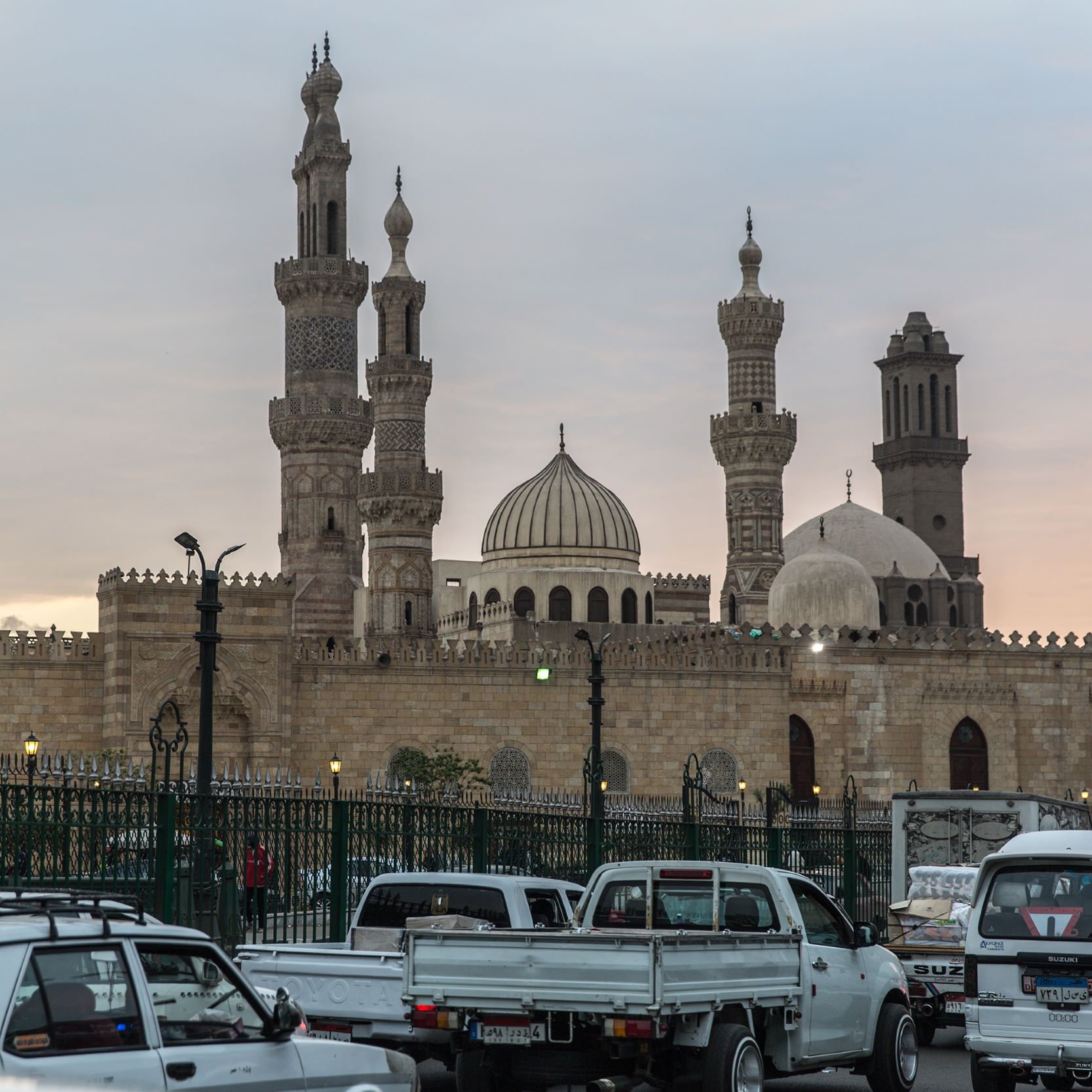 L'image montre un paysage urbain avec des bâtiments emblématiques, comprenant des mosquées avec des minarets élancés et des dômes caractéristiques. Au premier plan, on peut voir des véhicules traversant une route animée, entourés d'une clôture verte. L'éclairage doux de la fin de journée ajoute une atmosphère chaleureuse à la scène, tandis que le ciel présente des teintes nuancées, suggérant le coucher du soleil. L'architecture évoque un style traditionnel, typique de certaines grandes villes historiques.
