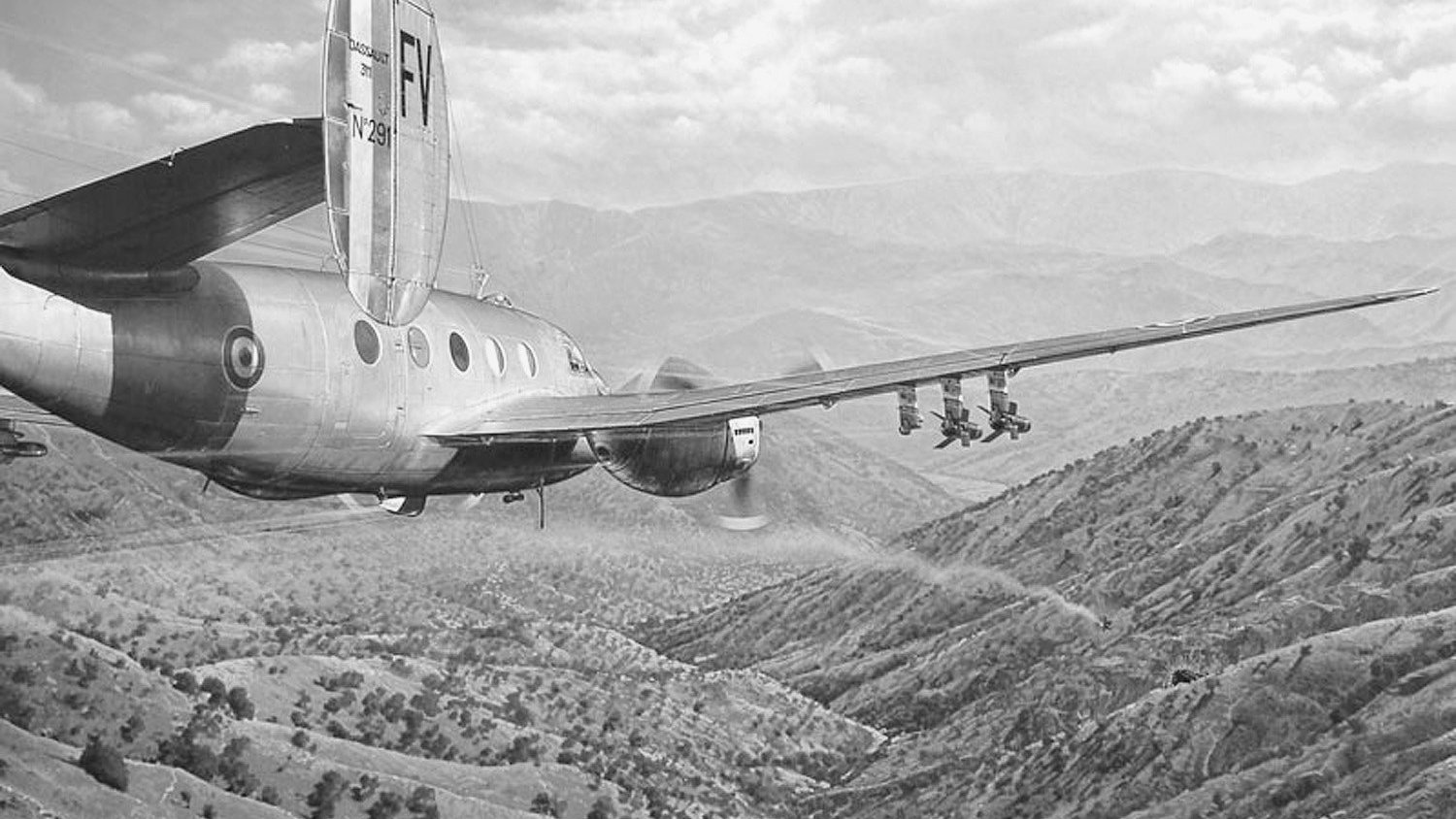 L'image montre un avion en vol, survolant un paysage montagneux. On peut voir des collines verdoyantes et des vallées en dessous, avec une atmosphère dramatique créée par les nuages. L'avion a un design classique, avec une queue distinctive et plusieurs réacteurs visibles. Ce type de scène évoque souvent des thèmes liés à l'aviation ou à la surveillance aérienne.