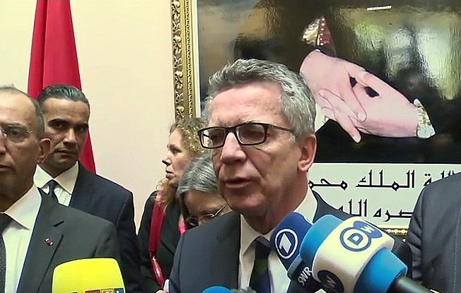 L'image montre un groupe de personnes lors d'une conférence de presse ou d'un événement officiel. Au premier plan, un homme s'exprime devant des caméras et des micros, entouré de plusieurs autres individus qui semblent attentifs. En arrière-plan, on peut voir un portrait ou une image représentant des mains avec des symboles significatifs, ainsi qu'un drapeau. L'atmosphère semble formelle, probablement liée à des discussions d'ordre politique ou diplomatique.