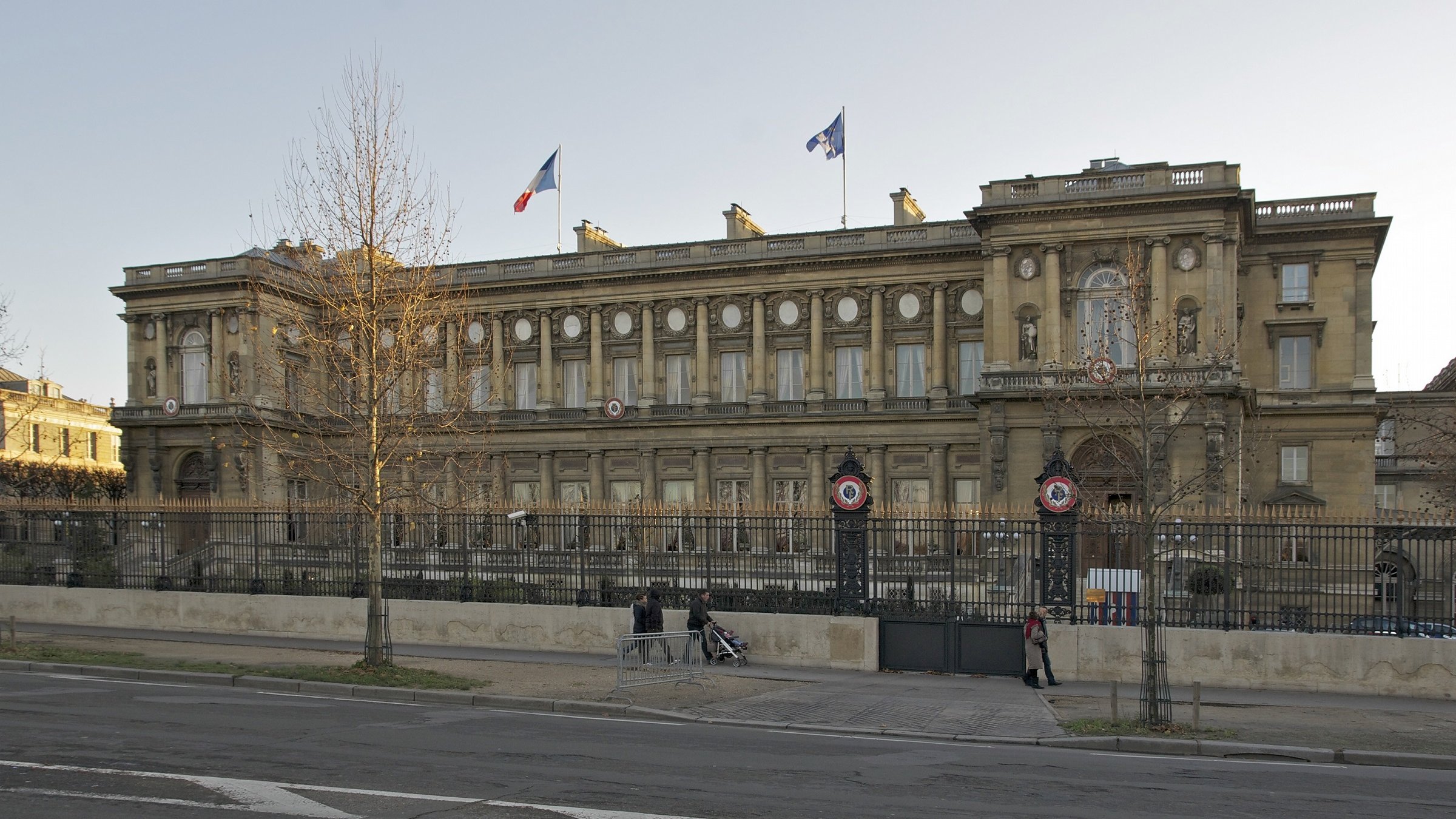L'image montre un bâtiment imposant avec une architecture classique, situé dans un espace urbain. On peut voir des drapeaux français flottant au sommet, et le bâtiment est entouré d'une grille. La façade est riche en détails, avec des colonnes et des ornements décoratifs. Il semble être un édifice gouvernemental ou historique, situé probablement dans une grande ville française. Des arbres sont visibles sur les côtés, et une route passe devant le bâtiment.