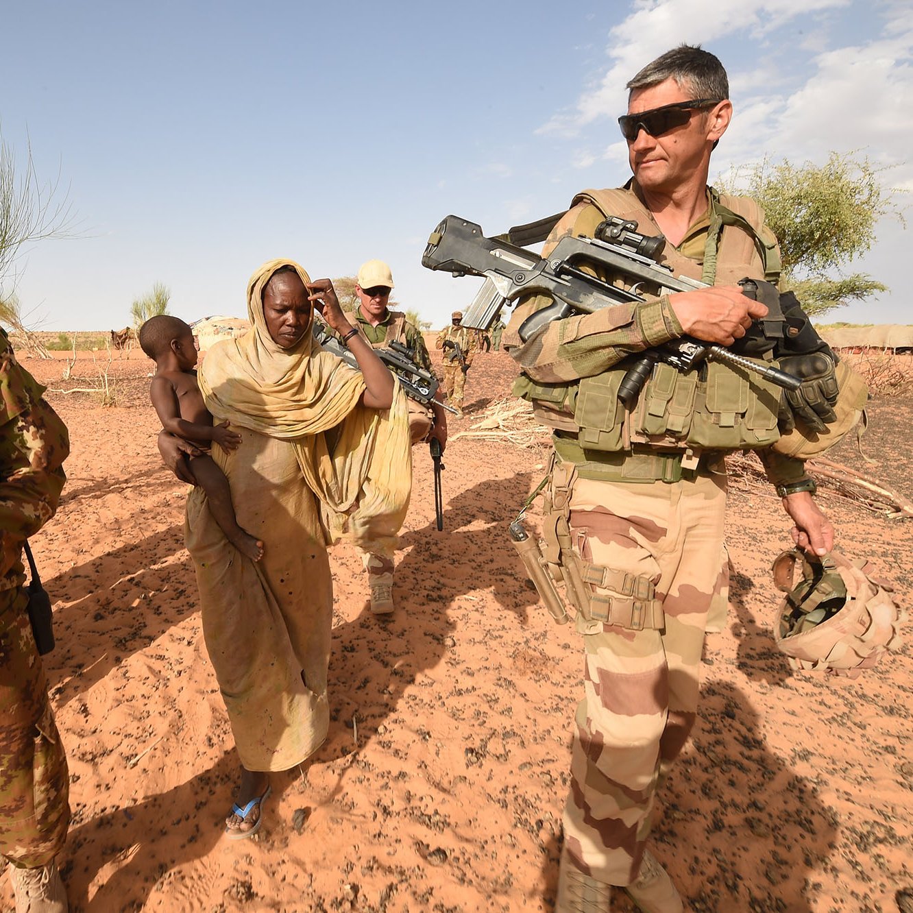The image shows a scene in a desert setting, featuring military personnel and civilians. On the left, a soldier in a camouflage uniform holds an automatic weapon and wears a helmet, while next to him, a woman in traditional attire carries a young child. To the right, another soldier, dressed in a different military uniform and also armed, is observing the surroundings. The background features sparse vegetation typical of a desert environment. The overall atmosphere suggests a military or peacekeeping operation, possibly in a humanitarian context.