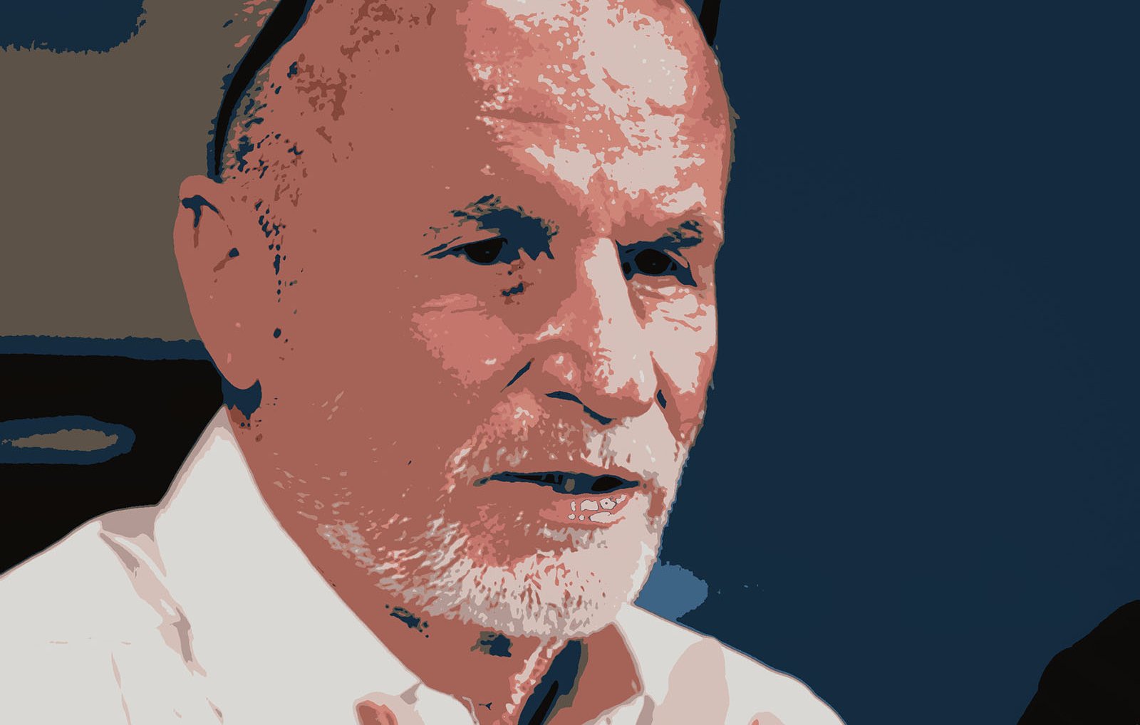 L'image représente un homme avec des cheveux grisonnants et une barbe. Il porte une chemise blanche et des lunettes posées sur sa tête. Son expression semble sérieuse et il a l'air de parler ou d'écouter attentivement. Le fond est de couleur sombre, ce qui met en valeur son visage.