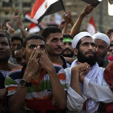 L'image montre un groupe de personnes rassemblées lors d'une manifestation. On peut voir des hommes de différentes origines, certains portant des vêtements traditionnels, d'autres des t-shirts colorés. Ils semblent tous attentifs et engagés, certains levant les mains ou tenant des drapeaux. L'ambiance semble être celle d'une mobilisation ou d'une expression de soutien pour une cause. Le fond montre un environnement urbain, avec d'autres manifestants et des drapeaux visibles.
