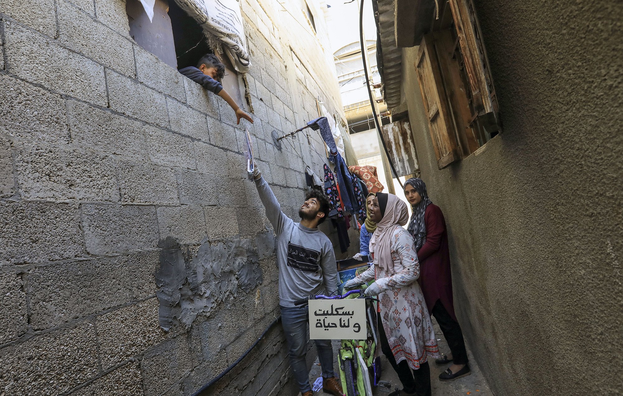 الصورة تُظهر مشهداً في زقاق ضيق حيث يبدو شاب يحمل شيئاً بيده وهو يمده نحو طفل يطل من نافذة في الجدار. في الجهة الأخرى، توجد امرأة ترتدي حجاباً متواجدة بجانب امرأة أخرى، وكلاهما تراقبان الموقف. يمكن رؤية أشياء معلقة في الجوار، مثل الملابس، وهناك دراجة ملونة موضوعة بجانب الجدار. الأجواء تعكس الحياة اليومية في منطقة مكتظة.