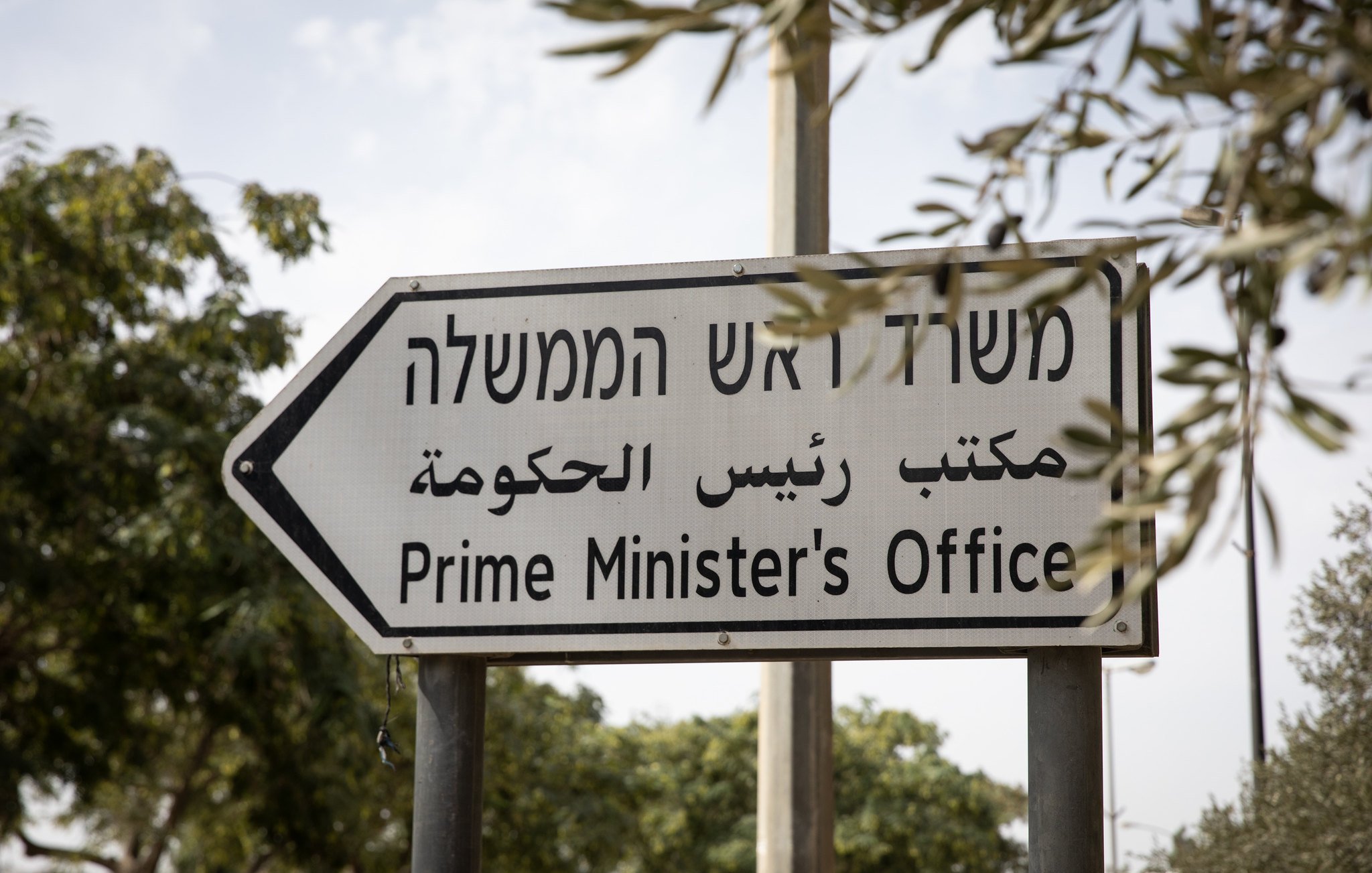 این تصویر یک تابلو راهنما را نشان می‌دهد که به زبان‌های عبری و عربی و انگلیسی نوشته شده است. متن تابلو به معنی "دفتر نخست‌وزیر" است. تابلو در یک محیط بیرونی قرار دارد و اطراف آن درختان و فضای سبز دیده می‌شود. طراحی تابلو ساده و واضح است و به نظر می‌رسد با هدف راهنمایی مردم به محل دفتر نخست‌وزیر نصب شده باشد.