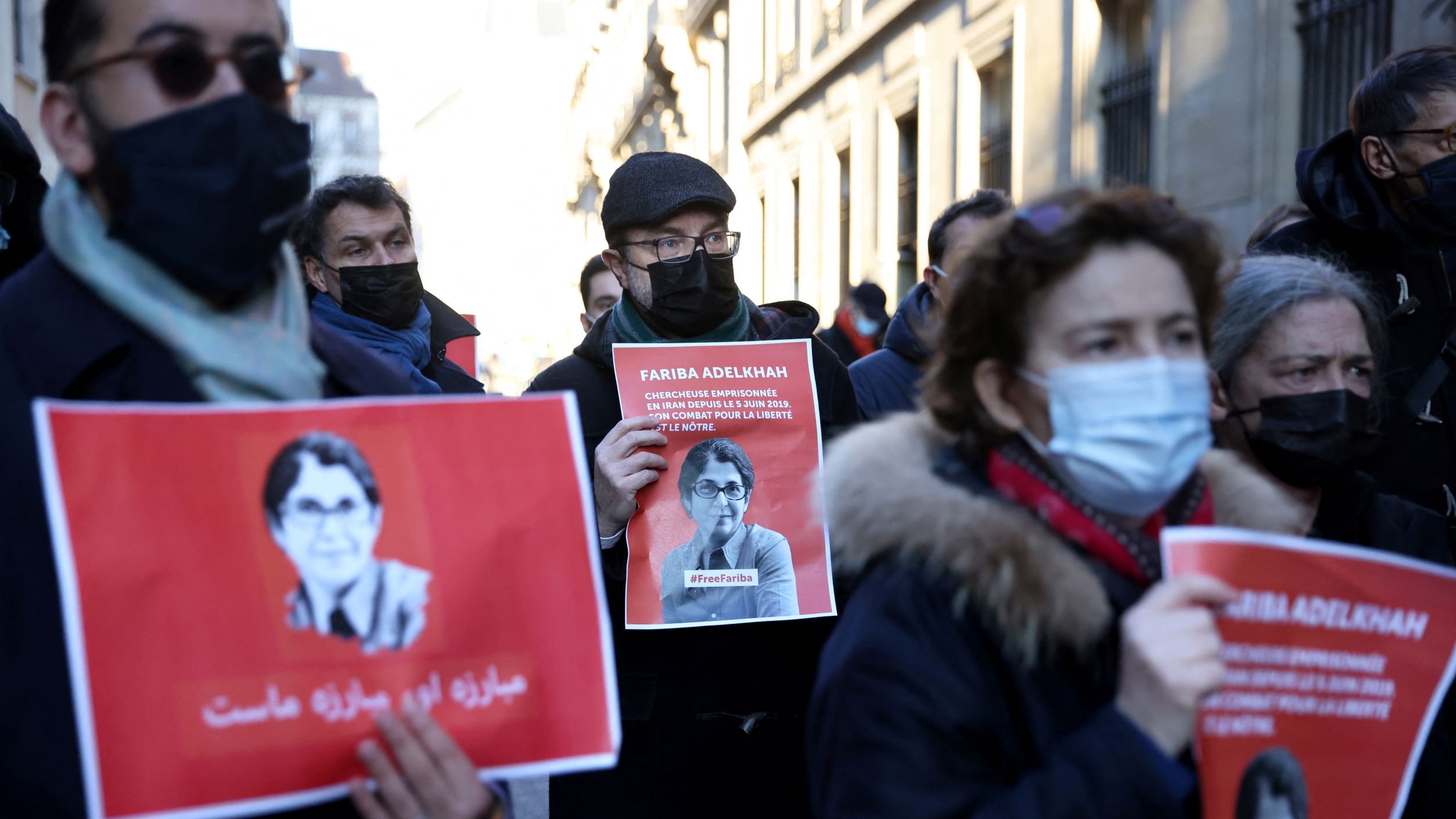 L'immagine mostra un gruppo di persone che partecipano a una manifestazione. Alcuni manifestanti tengono in mano cartelli con l'immagine e il nome di una persona, accompagnati da slogan scritti in arabo e in italiano. La maggior parte delle persone indossa mascherine e abbigliamento invernale, suggerendo che l'evento si svolge in un periodo freddo. L'atmosfera sembra essere seria e determinata, con un focus sulla questione rappresentata dai cartelli.