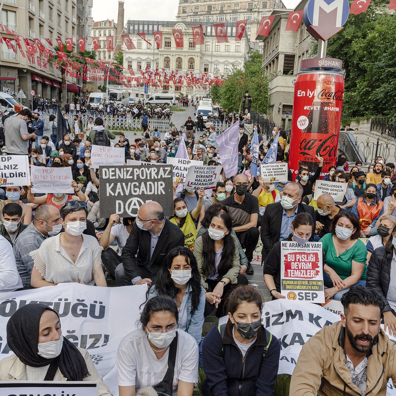 The image depicts a large gathering of people participating in a protest. Many of the individuals are wearing masks and holding various signs and banners with slogans, indicating their support for a cause. The scene appears to be in an urban setting, likely a city center, with flags in the background. The participants seem to be sitting on the ground, emphasizing their determination and solidarity for the issue they are advocating. The atmosphere looks charged with activism and communal spirit.