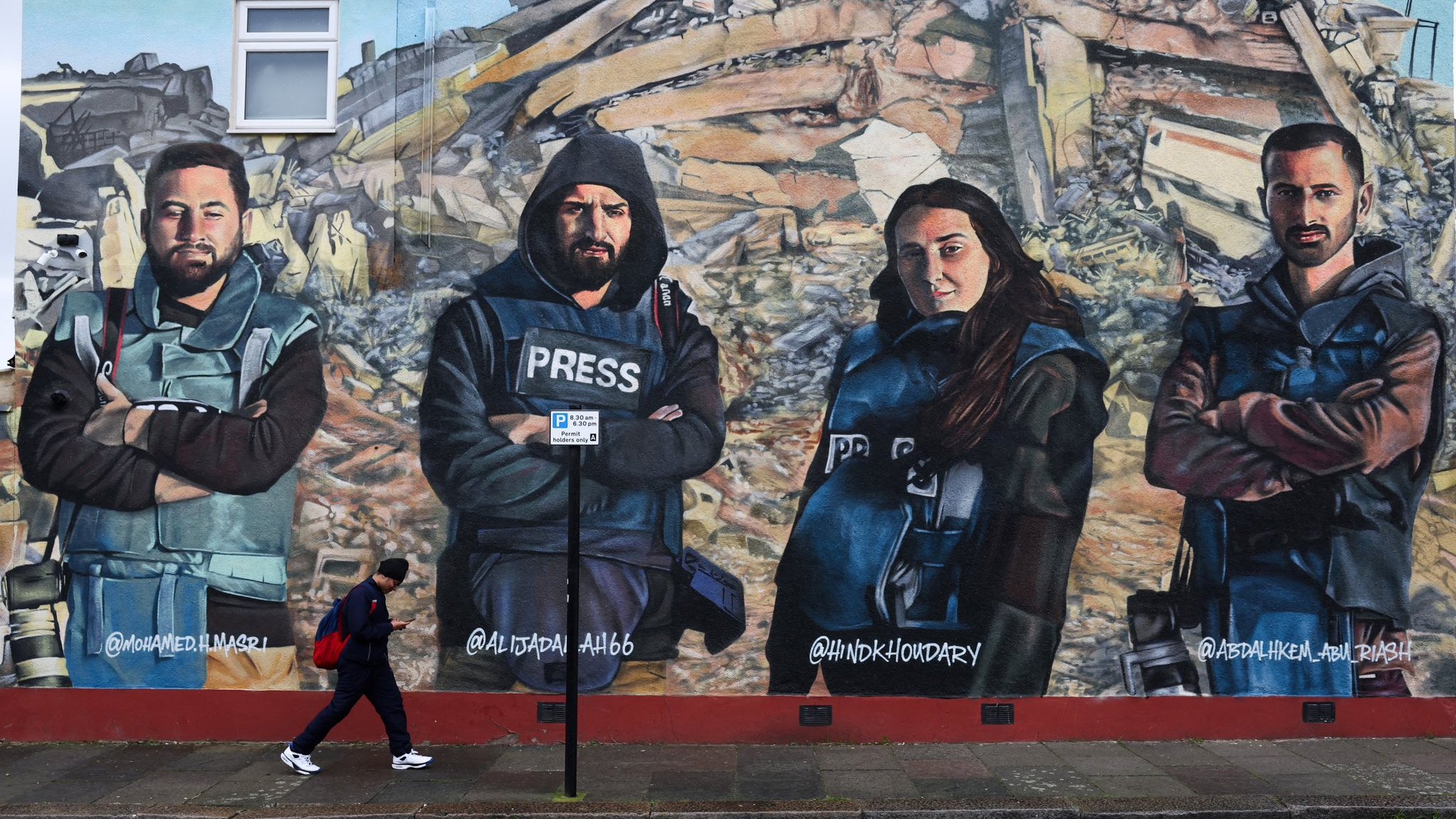 L'image montre une fresque murale représentant quatre personnes habillées en gilets pressés, posant avec les bras croisés. Ils semblent symboliser des journalistes ou des reporters. En arrière-plan, il y a des débris, suggérant un contexte de destruction ou de conflit. Une personne marche au premier plan, ajoutant une dynamique à la scène. Les visages des individus sur la fresque sont sérieux et déterminés.