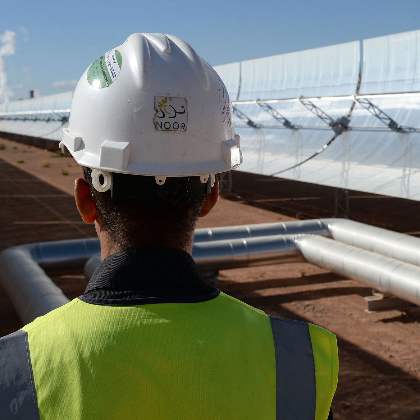 L'image montre un homme portant un casque de sécurité et un gilet de chantier, tourné vers un champ de miroirs solaires. Les miroirs sont orientés vers le soleil et semblent faire partie d'une installation de production d'énergie solaire. À l'arrière-plan, on peut apercevoir des tuyaux métalliques qui transportent probablement un fluide caloporteur. L'environnement est désertique, avec un ciel bleu dégagé qui suggère des conditions solaires idéales.