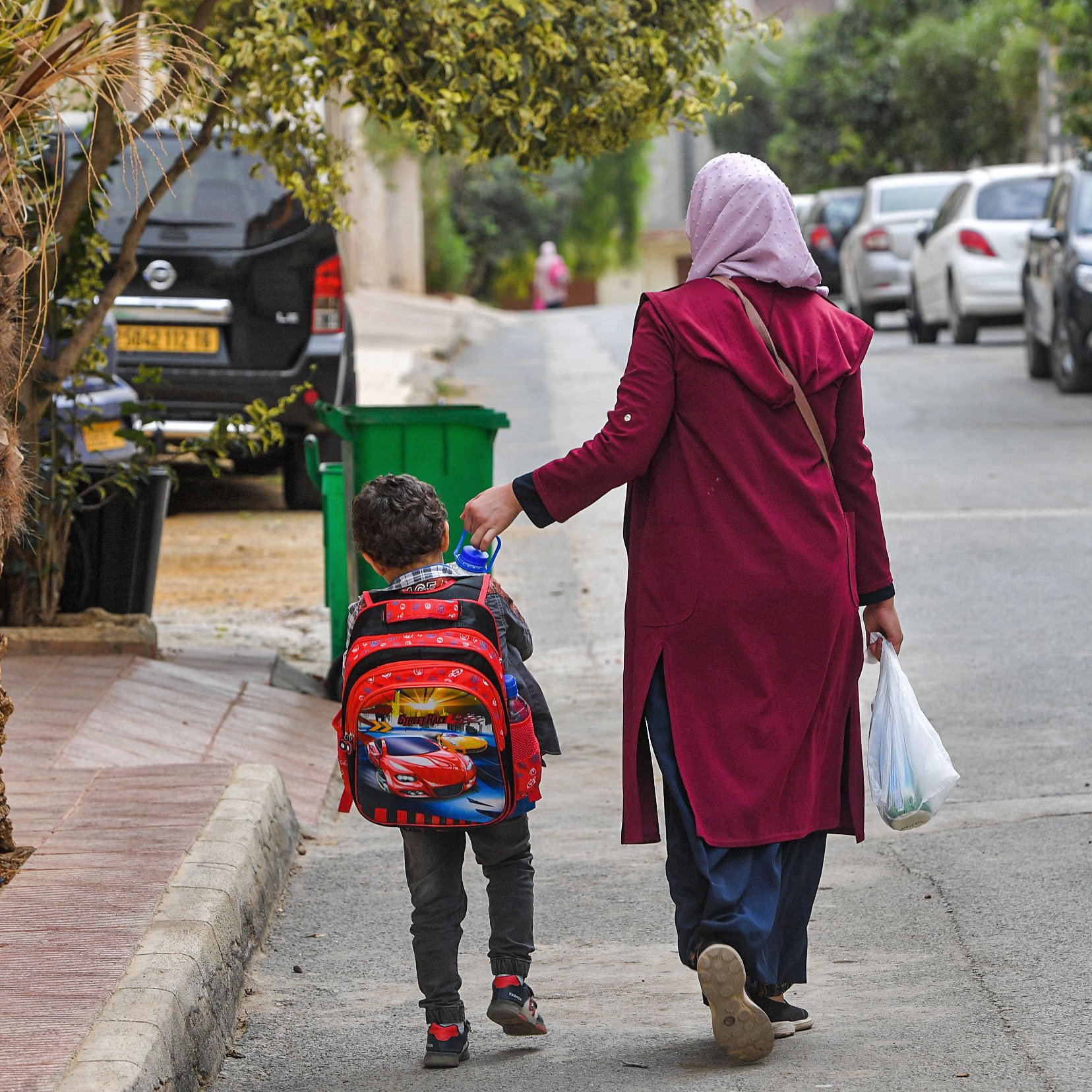 L'image montre une femme portant un foulard, vêtue d'un long manteau, qui marche dans une rue résidentielle avec un jeune garçon. Le garçon porte un sac à dos coloré, probablement inspiré d'un personnage de dessin animé. Ils semblent se diriger vers une destination, et la femme tient également un sac en plastique. En arrière-plan, on peut voir des voitures garées des deux côtés de la rue et de la verdure autour. L'atmosphère semble calme et quotidienne.