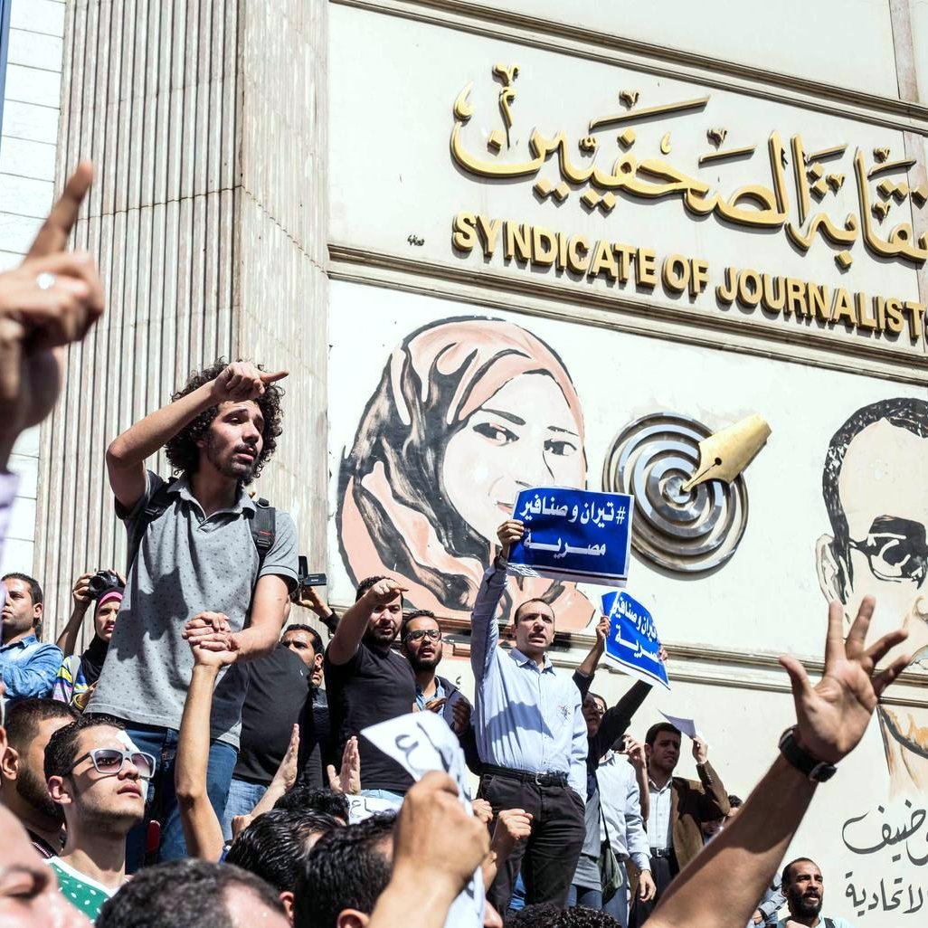 L'image montre une scène de manifestation devant un bâtiment, probablement le Syndicat des journalistes. De nombreuses personnes sont rassemblées, levant les bras en signe de protestation. Certains tiennent des pancartes ou des banderoles, tandis qu'une personne debout sur une foule semble s'adresser aux manifestants. L'arrière-plan présente une grande fresque représentant des figures, ajoutant à l'impact visuel de la scène. L'atmosphère est celle d'un rassemblement engagé et passionné.