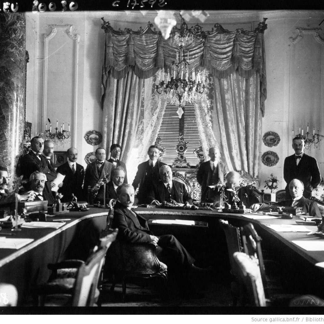 L'image montre une grande réunion dans une salle ornée. Au centre, plusieurs hommes sont assis autour d'une grande table, tandis que d'autres se tiennent debout en arrière-plan. La pièce est décorée avec des rideaux élégants et des éléments architecturaux raffinés, tels que de grands lustres et des colonnes. Les participants semblent engagés dans une discussion sérieuse, reflétant l'importance de l'événement. L'atmosphère est formelle et historique.