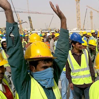 L'image montre un grand groupe de travailleurs, majoritairement en train de lever les bras. Tous portent des casques de sécurité jaunes et des gilets réfléchissants. L'environnement semble être un site de construction, avec plusieurs grues et équipements visibles en arrière-plan. L'atmosphère paraît dynamique et l'affluence des travailleurs indique probablement une activité importante sur le chantier.