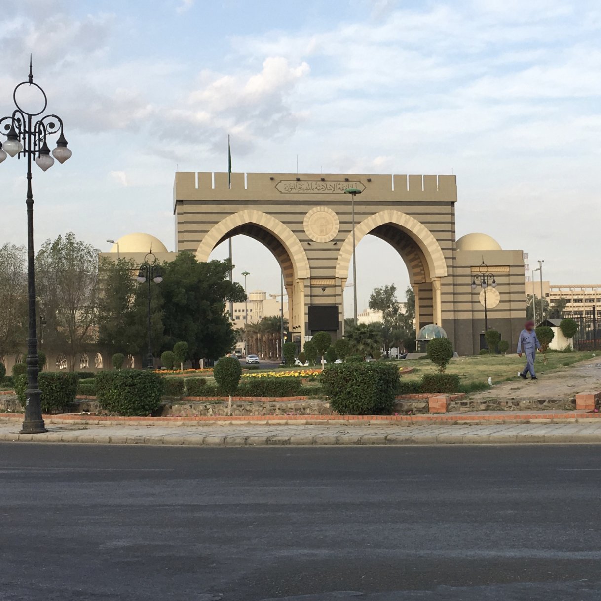 A grand archway with domes, surrounded by greenery and a road, under a cloudy sky.