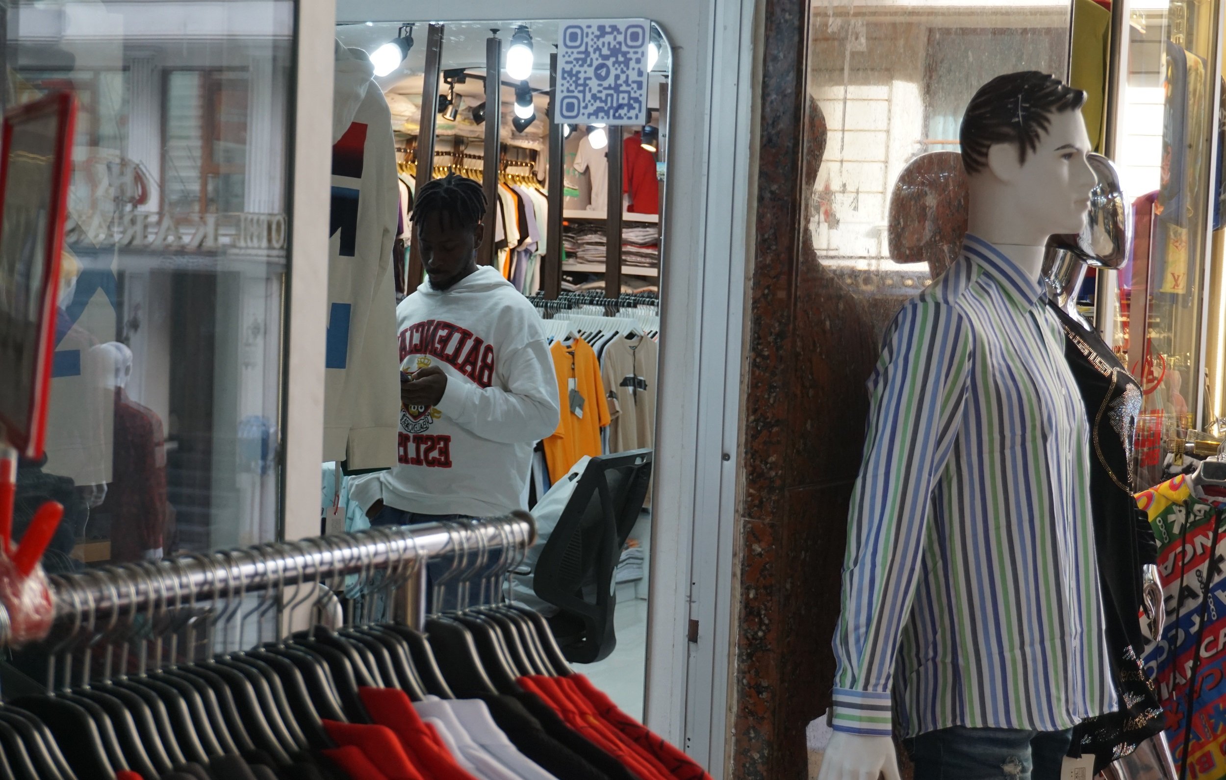 L'image montre un magasin de vêtements avec des rangées de vêtements accrochés, notamment des t-shirts et des chemises multicolores. À gauche, un homme portant un sweat-shirt blanc est en train de regarder son téléphone. À droite, un mannequin habillé d'une chemise à rayures est exposé dans la vitrine. On peut aussi apercevoir un code QR accroché à l'intérieur du magasin. L ambiance est typique d'un espace commercial animé.