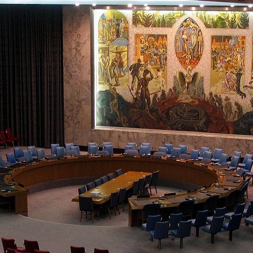 L'image montre l'intérieur d'une salle de réunion, probablement celle du Conseil de sécurité des Nations Unies. On y voit un grand panneau mural coloré représentant des scènes symboliques. La salle est circulaire avec une table ovale au centre, entourée de chaises disposées en arcs. Les chaises sont de différentes couleurs, majoritairement bleues et rouges. L'éclairage est doux, mettant en valeur les œuvres d'art sur le mur. L'atmosphère semble formelle et solennelle, propice aux discussions diplomatiques.