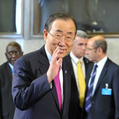 L'image montre un homme souriant, vêtu d'un costume sombre avec une cravate rose. Il semble faire un geste de la main, comme s'il saluait. En arrière-plan, on peut apercevoir d'autres hommes en costume, certains portant des badges. L'environnement semble être un lieu formel, probablement dans un contexte de réunion ou de conférence internationale.