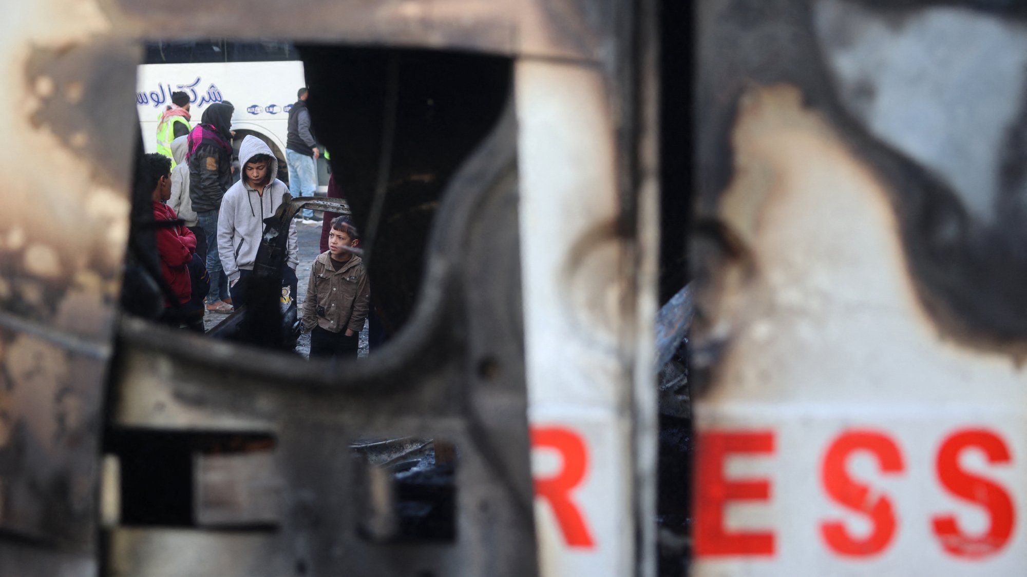 L'image montre un groupe de personnes, y compris des enfants, rassemblées dans un environnement urbain perturbé. On aperçoit une structure endommagée, peut-être un véhicule, qui présente des signes de destruction. Le reflet dans la structure suggère une atmosphère de tension ou de conflit, tandis que les visages des personnes expriment diverses émotions. L'environnement semble chaotique, soulignant une situation difficile.
