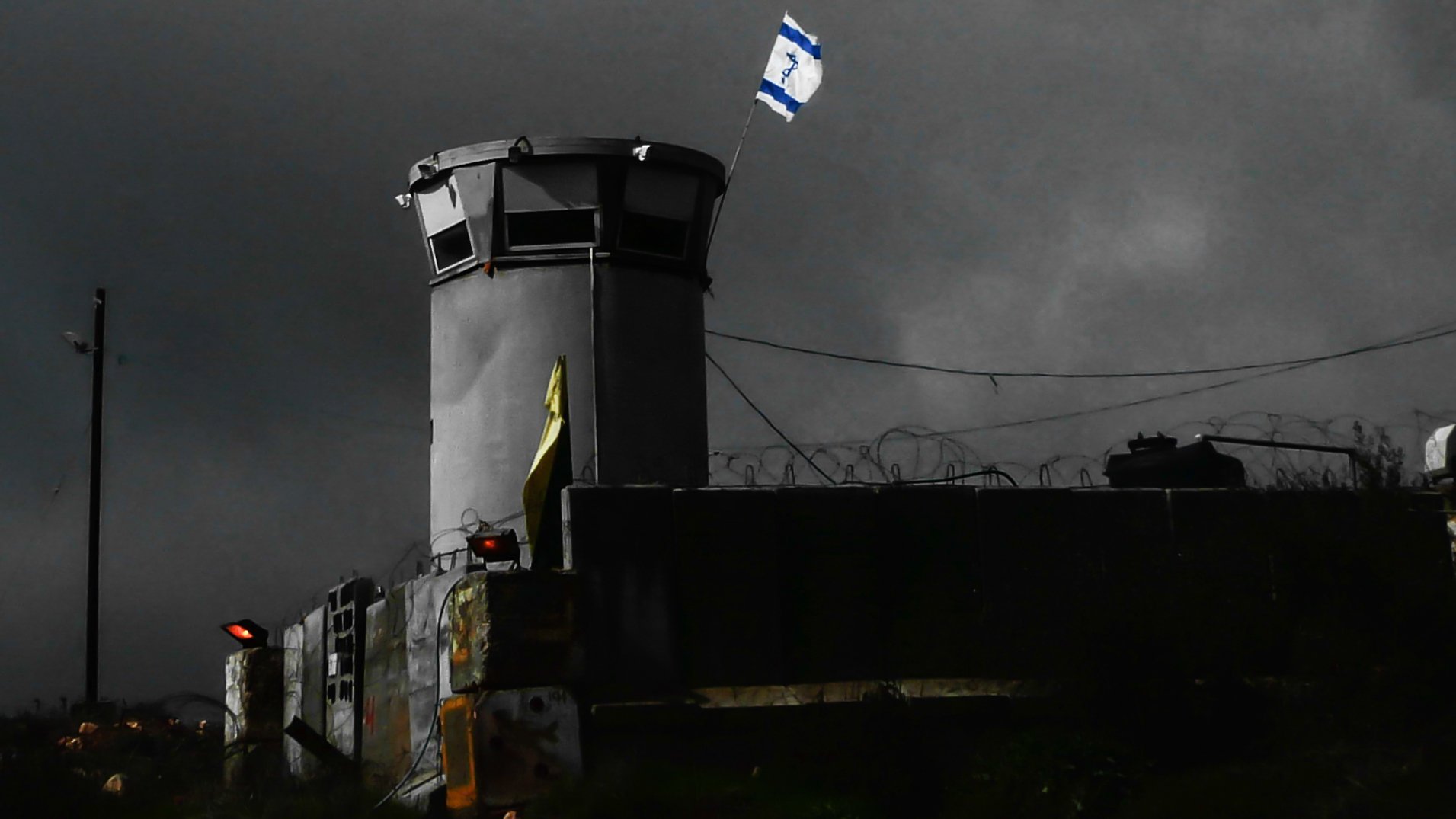 L'image montre une structure semblable à une torre de surveillance, située dans un environnement sombre et nuageux. On peut voir un drapeau israélien flottant au sommet de la tour. Des barbelés sont visibles autour du périmètre, ce qui suggère une zone sécurisée ou protégée. L'éclairage de la scène crée une atmosphère de tension, accentuant l'aspect austère du lieu.