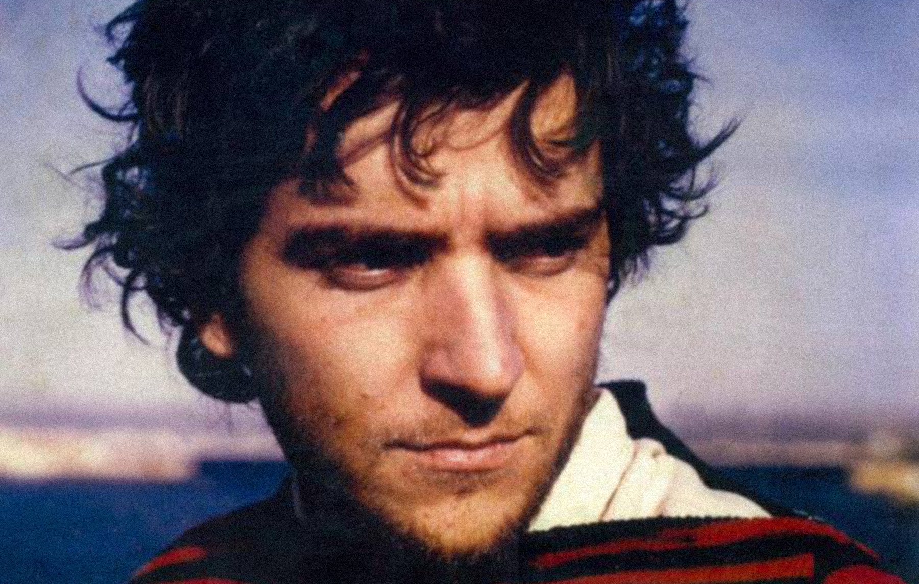 L'image montre un jeune homme avec des cheveux foncés et ondulés, portant un pull au motif rayé. Son expression semble pensive, et il regarde légèrement sur le côté. L'arrière-plan est flou, suggérant un paysage maritime ou naturel, ce qui ajoute une atmosphère contemplative à la scène. Les couleurs de l'image sont douces et légèrement nostalgiques.