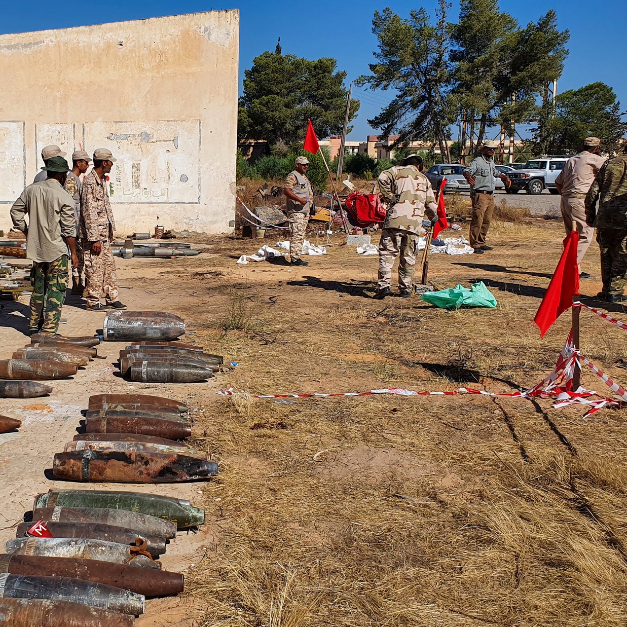تظهر الصورة مجموعة من الأشخاص يرتدون زيًا عسكريًا، يتواجدون في منطقة مفتوحة تحتوي على العديد من القذائف والذخائر القديمة المصفوفة على الأرض. الخلفية تحتوي على جدران مبنى متهدم، بينما تظهر شجيرات وأشجار في المنطقة المحيطة. يبدو أن هناك بعض الأعلام والأعلام الحمراء في الموقع، مما يشير إلى نشاطات أو إجراءات تتعلق بالسلامة أو التفكيك. الجو العام يوحي بأن هناك عملية تُجرى للتعامل مع هذه الذخائر.