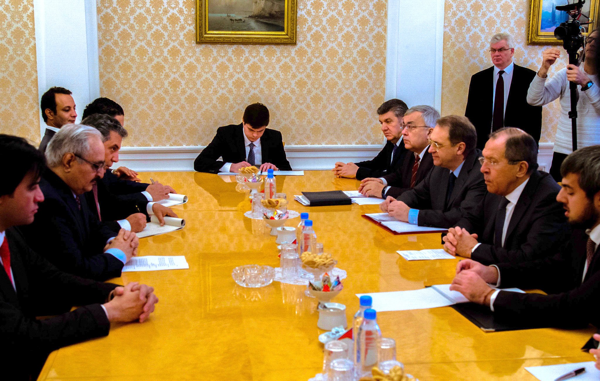 تظهر الصورة اجتماعاً رسمياً يتم فيه نقاش أو تبادل للأفكار بين مجموعة من الأشخاص. يجلس المشاركون حول طاولة كبيرة تحتوي على مستندات وبعض الوجبات الخفيفة. يبدو أن هناك تركيزاً على النقاش، حيث يظهر البعض وهم يكتبون أو يتطلعون إلى المستندات الموجودة أمامهم. الخلفية تحتوي على لوحات فنية تزين الجدران، مما يضيف طابعاً رسمياً إلى الاجتماع.