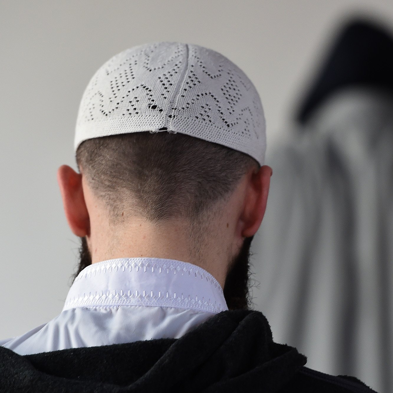 الصورة تظهر شخصًا يرتدي قبعة تقليدية بيضاء، مع ظهور جزء من رأسه وظهره. خلفه يوجد شخص آخر يرتدي غطاء رأس داكن مع معطف، مما يخلق تباينًا في الألوان. الجو العام للصورة يوحي بالهدوء والتركيز، وقد تكون في سياق ديني أو روحي.