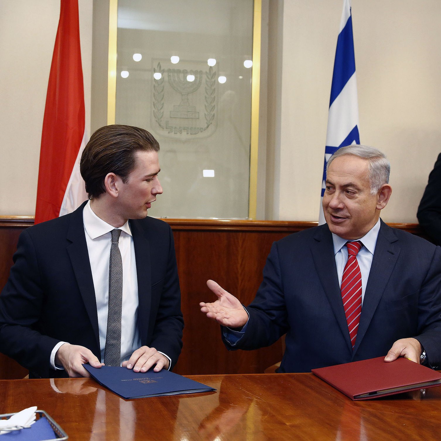 L'image montre deux hommes assis à une table, probablement lors d'une réunion ou d'une cérémonie de signature. L'un d'eux est en costume sombre avec une cravate claire, tandis que l'autre porte une cravate rouge. Ils semblent engagés dans une conversation amicale. À l'arrière-plan, on peut voir des drapeaux, probablement ceux de leurs pays respectifs. L'ambiance générale semble formelle et diplomatique.