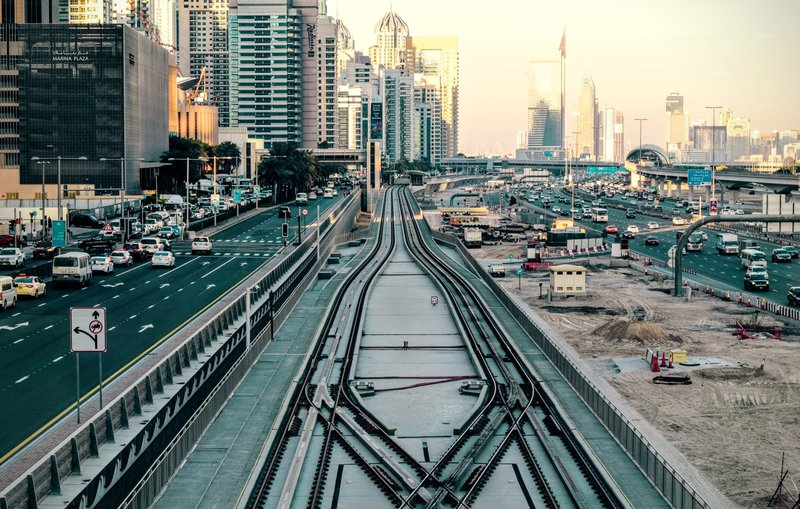 L'image montre une vue urbaine d'une ville moderne. On peut voir des routes animées avec des voitures circulant, ainsi qu'un important réseau de rails de train. À droite, il y a des bâtiments imposants et modernes qui reflètent l'architecture contemporaine. L'atmosphère est lumineuse, suggérant un moment de la journée proche du coucher du soleil avec des lumières douces. Les rails semblent bifurquer, suggérant une intersection, tandis que des travaux de construction ou d'aménagement sont visibles au premier plan.