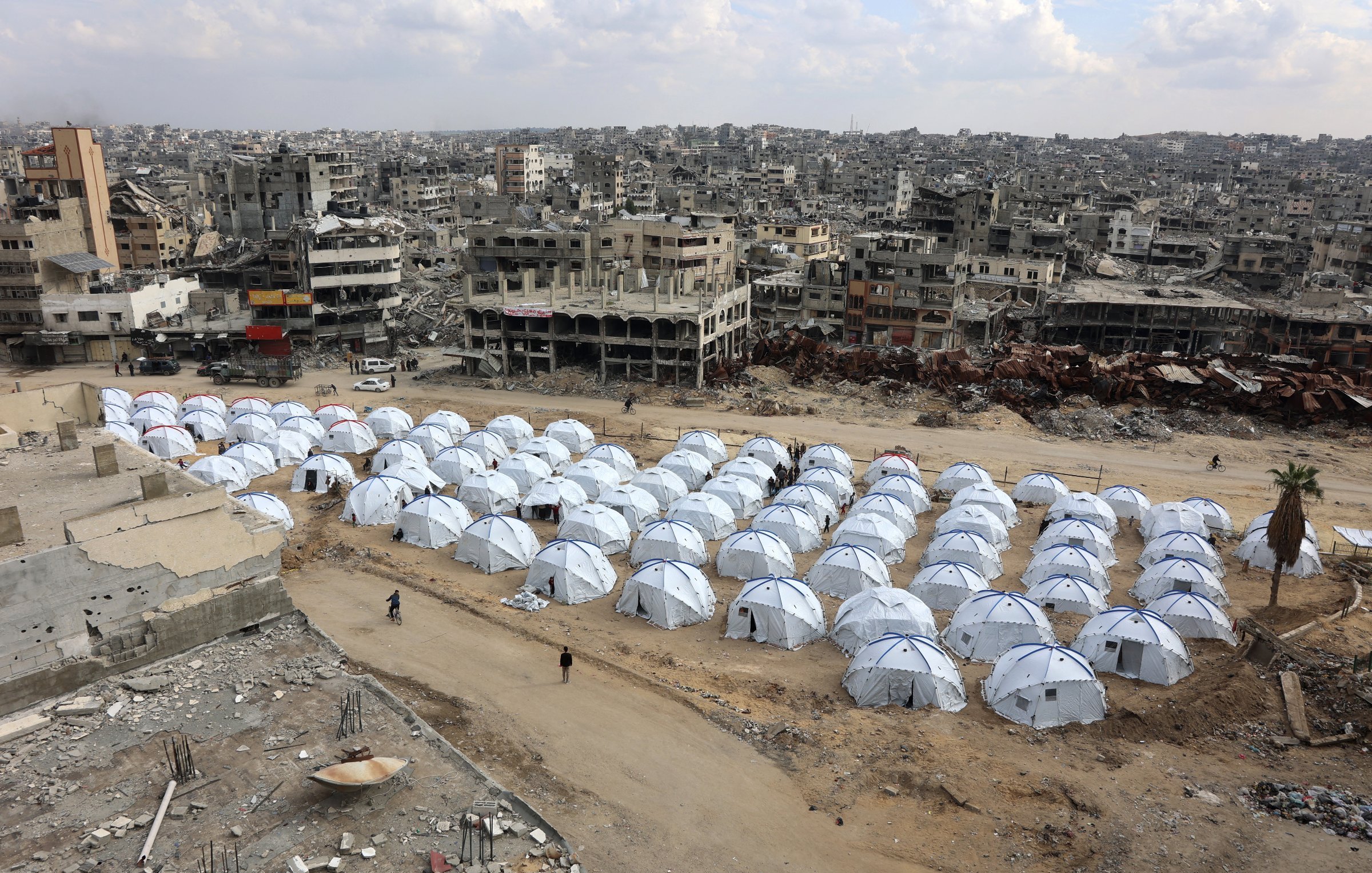 این تصویر نمایی از یک منطقه تخریب‌شده را نشان می‌دهد که در آن چادرهای سفید رنگ به‌عنوان محل اسکان موقت قرار گرفته‌اند. در پشت این چادرها، ساختمان‌های ویران دیده می‌شوند که نشان‌دهنده آثار جنگ یا بلایای طبیعی هستند. زمین خاکی و خالی با چندین چادر در صف‌های منظم، حس نیاز و بحران را منتقل می‌کند. آسمان نیز ابری به نظر می‌رسد که به فضای تصویر افزوده است.