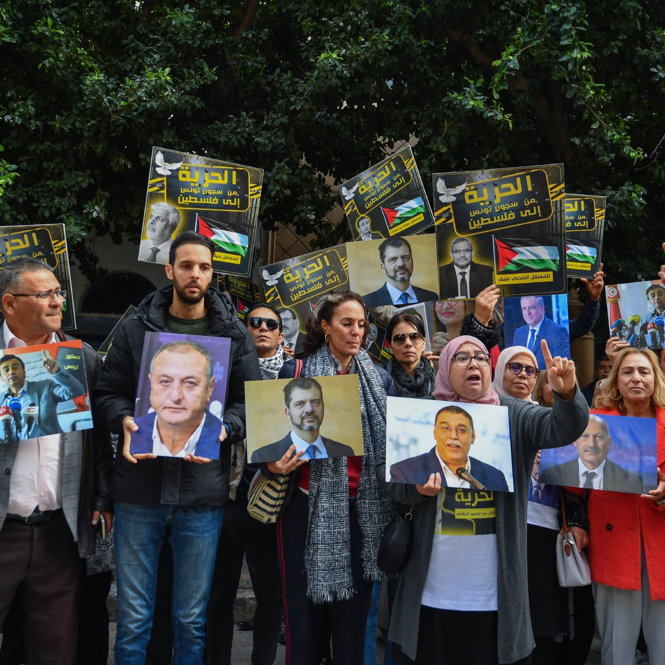 L'image montre un groupe de personnes réunies lors d'une manifestation. Elles tiennent des pancartes et des portraits de personnalités, exprimant des messages de soutien. L'ambiance est dynamique, avec des visages souriants et engagés.