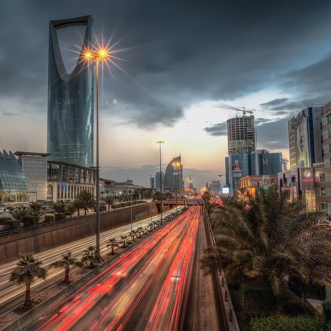 The image depicts a bustling urban scene, likely in a modern city. In the foreground, streaks of light from moving vehicles indicate heavy traffic on a multi-lane road, while palm trees line the sides, adding a touch of greenery. In the background, prominent skyscrapers with reflective glass facades dominate the skyline under a cloudy sky, creating a dramatic atmosphere. The scene captures a blend of modern architecture and vibrant city life, illuminated by streetlights and the fading light of sunset.
