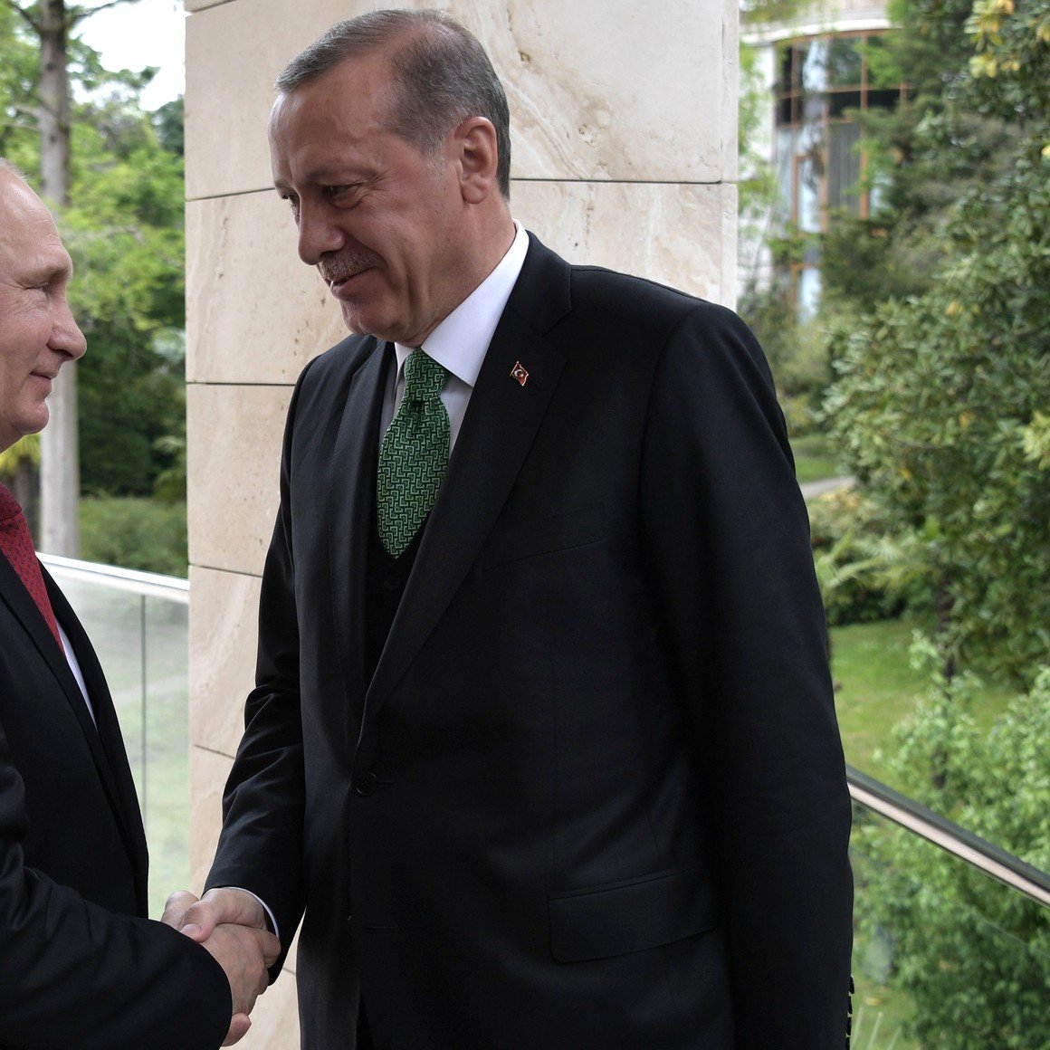 در این تصویر، دو شخصیت سیاسی برجسته در حال دست دادن هستند. یکی از آن‌ها مردی با موهای کوتاه و رنگ طلایی، دارای کت و شلوار سیاه و پیراهن سفید است. دیگری مردی با موهای تیره و کت و شلوار تیره و کروات سبز. زمینه تصویر شامل سبزیجات و فضای طبیعی است که نشان‌دهنده یک محیط آرام و دوستانه می‌باشد. هر دو شخصیت ظاهر جدی دارند و در حال تبادل گفت‌وگو هستند.