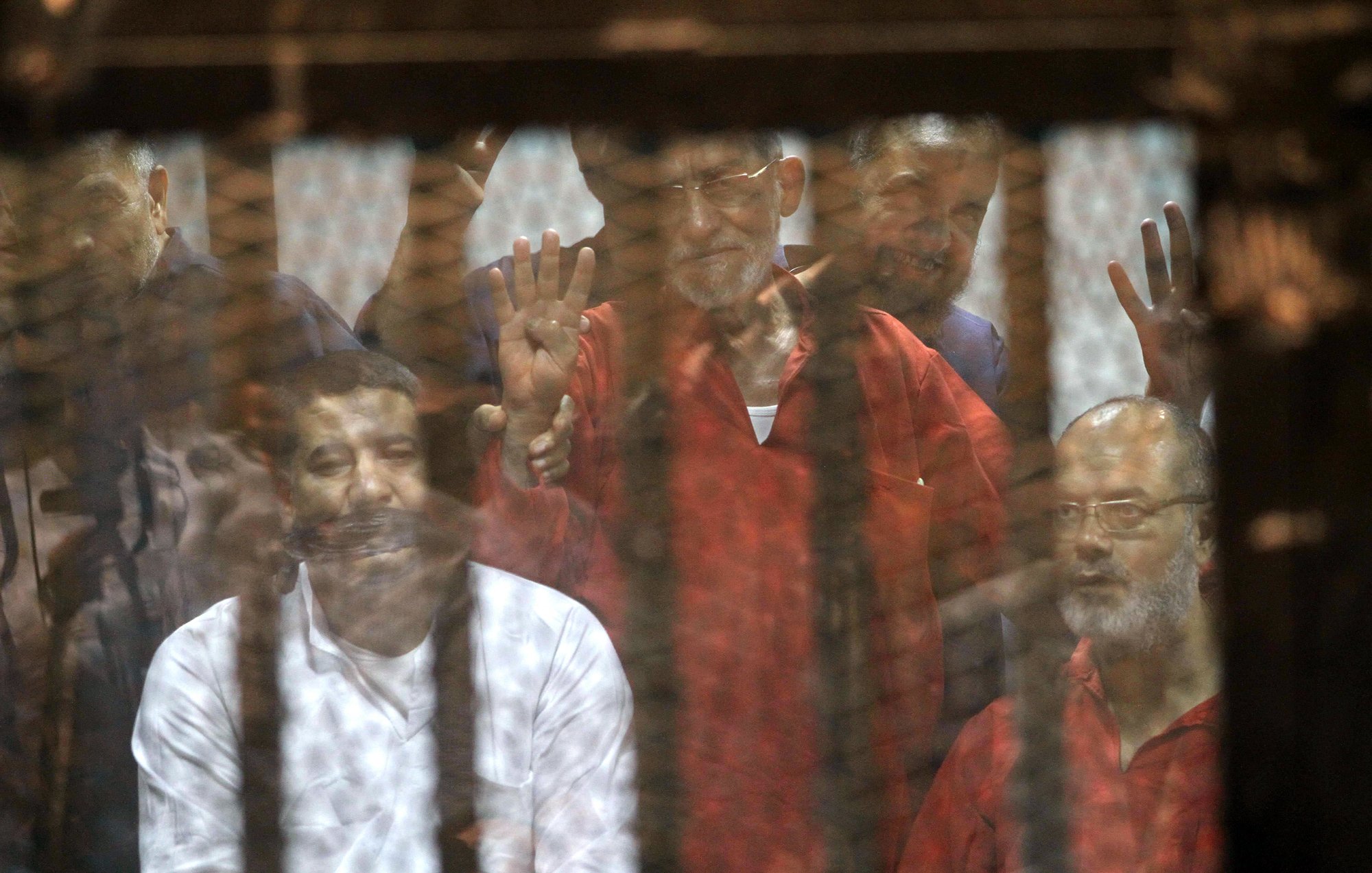 L'image montre un groupe d'hommes derrière une grille ou un panneau de verre. Ils semblent être dans un contexte de détention ou de procès, avec des expressions variées sur leurs visages. Certains lèvent les mains, suggérant un geste de protestation ou de solidarité. L'ambiance générale est sombre, évoquant des thèmes de justice et de droits humains.