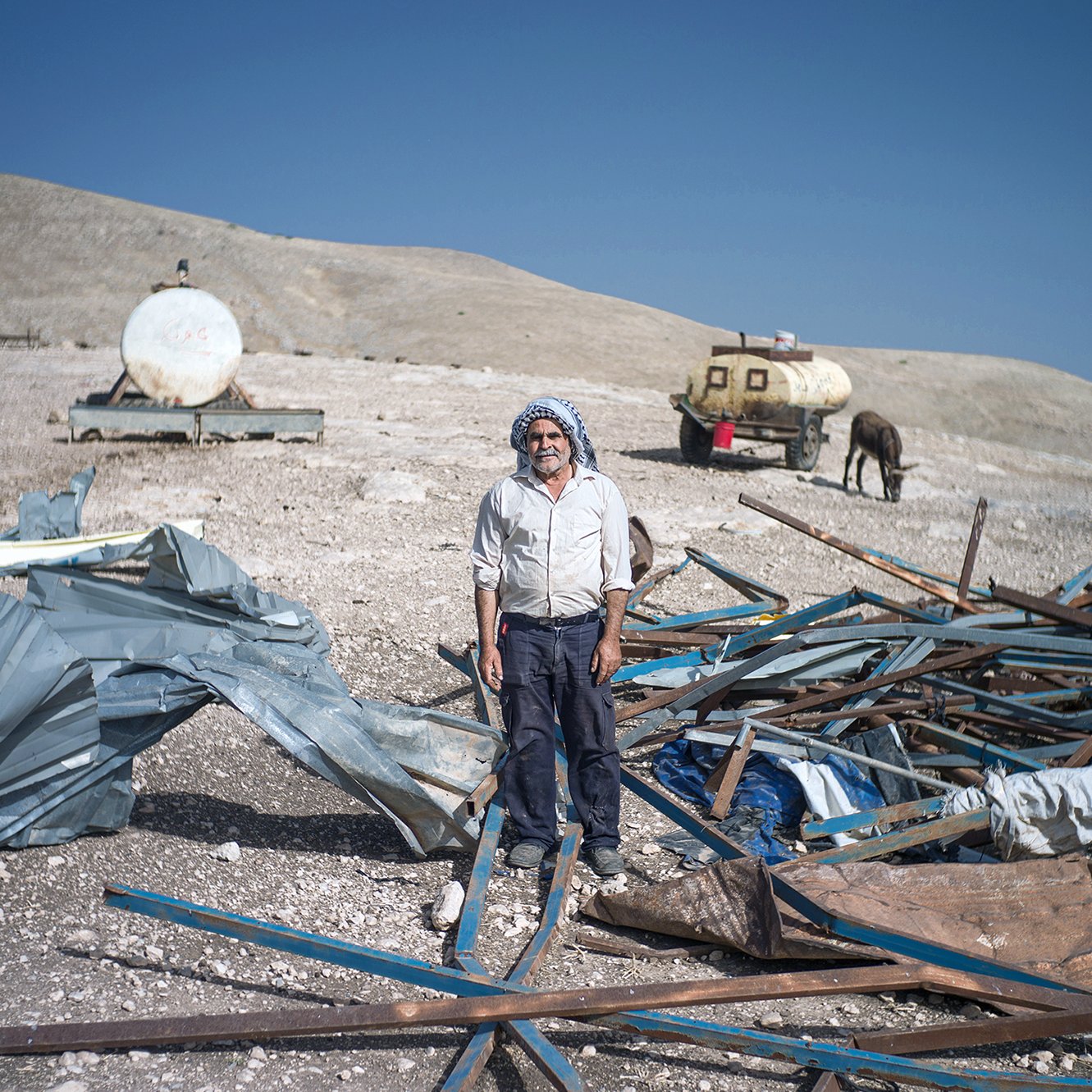 L'image montre un homme debout au milieu d'un terrain désolé. Il est entouré de débris, comprenant des structures métalliques et des morceaux de toile. En arrière-plan, on peut voir des réservoirs ou des citernes, ainsi qu'un cheval. Le paysage semble aride, avec des collines en arrière-plan sous un ciel bleu. L'homme porte une tenue simple et dégage une impression de résilience dans un environnement difficile.