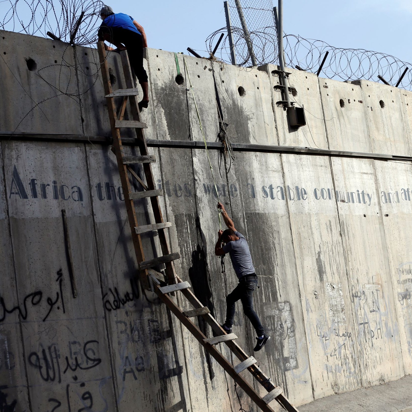 L'image montre une scène où deux personnes tentent de monter ou de descendre d'un mur de béton. Une échelle est appuyée contre le mur, et il y a des barbelés au sommet. Le mur présente également des graffitis ou des inscriptions, et l'environnement semble urbain. Les personnes semblent concentrées sur leur tâche, soulignant une dynamique d'effort et de défi.