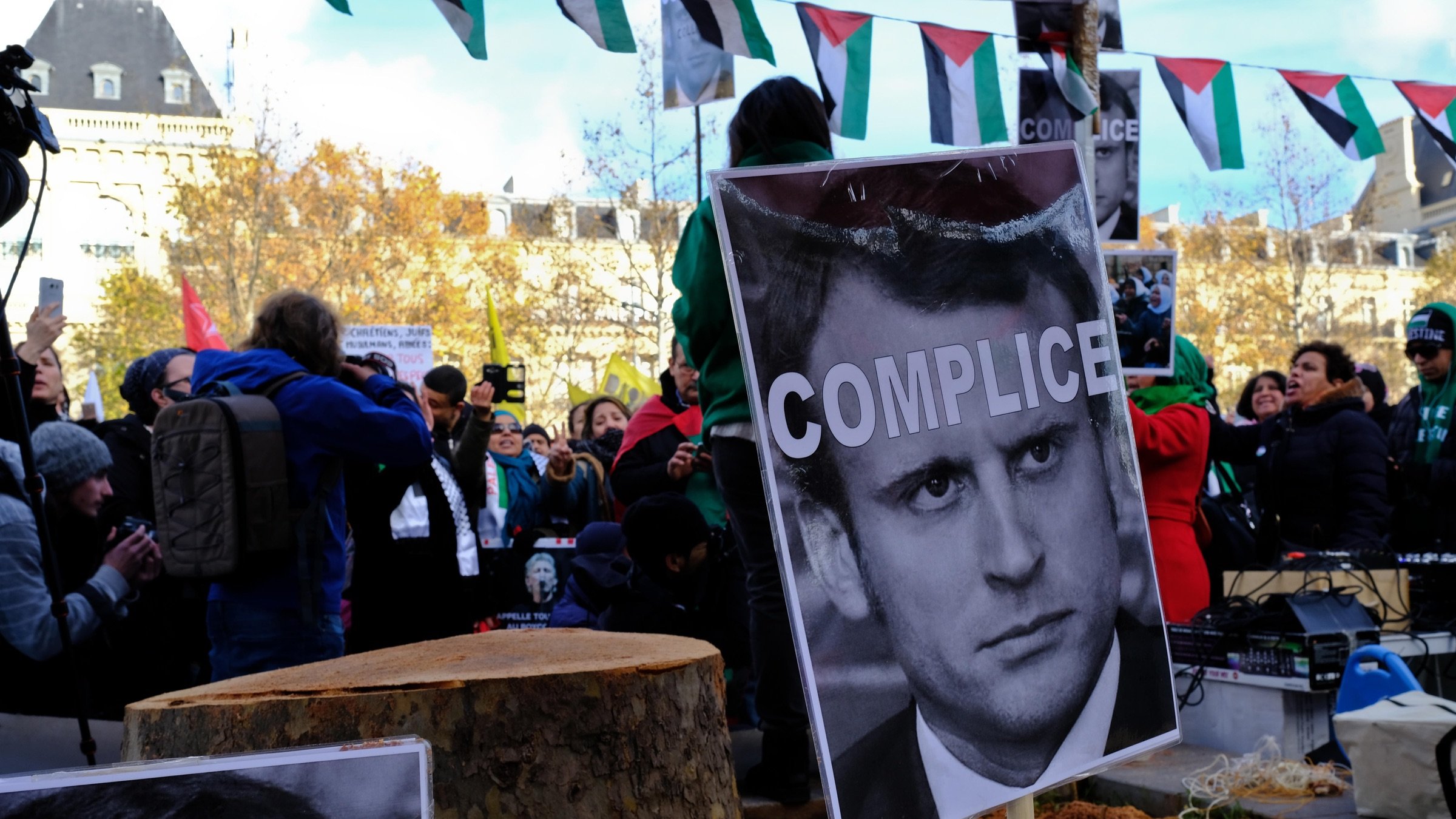 L'image montre une manifestation où une foule se rassemble autour d'un tronc d'arbre coupé. Des panneaux sont affichés avec des portraits et le mot "COMPLICE" écrit en gros caractères sur une affiche. En arrière-plan, on peut voir des drapeaux et des personnes prenant part à la protestation. L'atmosphère semble tendue, illustrant des sentiments de mécontentement et des revendications politiques.