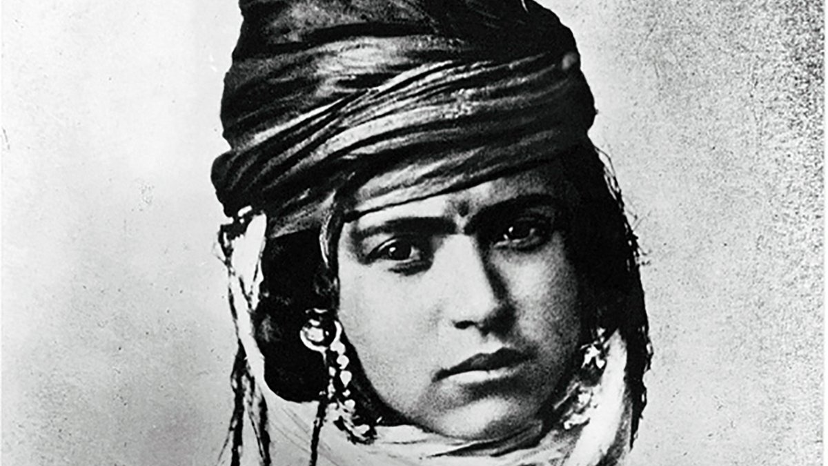 L'image montre un portrait noir et blanc d'une personne portant un turban orné et des bijoux. Le regard de la personne est intense, et son expression transmet une certaine dignité. Elle est vêtue de vêtements traditionnels qui reflètent une culture spécifique. Les détails du turban et des ornements ajoutent une dimension riche et texturée à l'image.