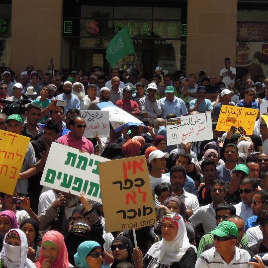 L'image montre une grande foule rassemblée lors d'une manifestation. Les participants tiennent divers panneaux et affiches, certains portant des messages en plusieurs langues, y compris l'hébreu et l'arabe. L'atmosphère semble dynamique, avec des gens exprimant leurs opinions et leurs préoccupations. On peut voir des drapeaux et des bannières, indiquant un fort engagement communautaire et un appel à l'action.