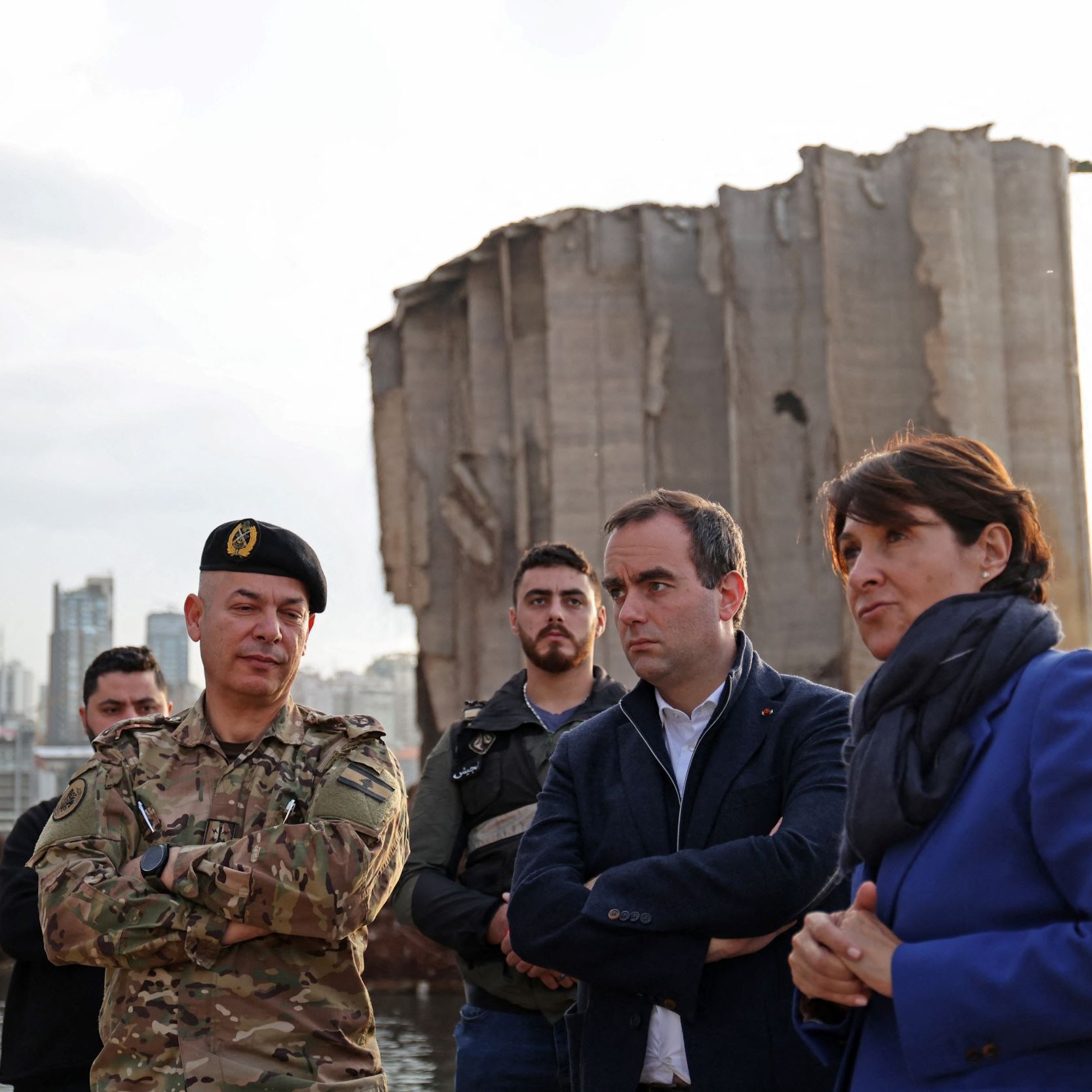 تظهر في الصورة مجموعة من الأشخاص في منطقة تضررت من انفجار أو حدث كارثي. في المقدمة، يوجد ضابط عسكري يرتدي زيًا رسميًا ولديه شارة على القبعة. بجانبه، يقف مجموعة من الرجال والنساء يتحدثون بجدية، مع تعابير وجه تعكس الانتباه والتفكير. في الخلفية، توجد آثار دمار واضحة، مع مبنى شاهق متضرر بشكل كبير. السماء تميل إلى اللون الرمادي، مما يضفي أجواءً من الحزن أو التأمل على المشهد.