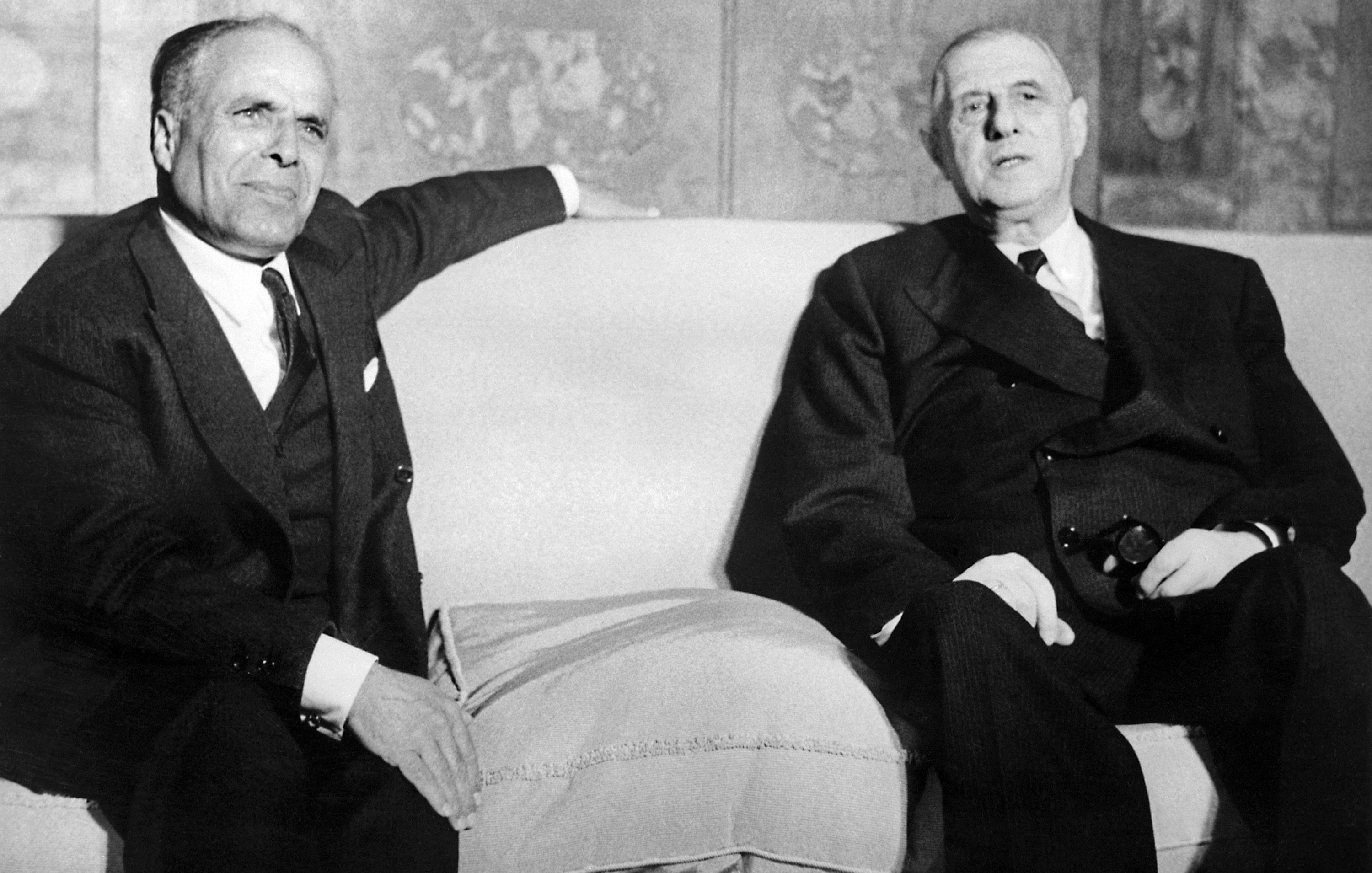 L'image montre deux hommes assis sur un canapé dans un environnement formel. Ils ont l'air sérieux et portent des costumes sombres. L'un des hommes semble légèrement se pencher en arrière, tandis que l'autre regarde devant lui. L'arrière-plan est décoré, mais les détails ne sont pas clairement visibles. L'atmosphère générale apparaît académique et solennelle.