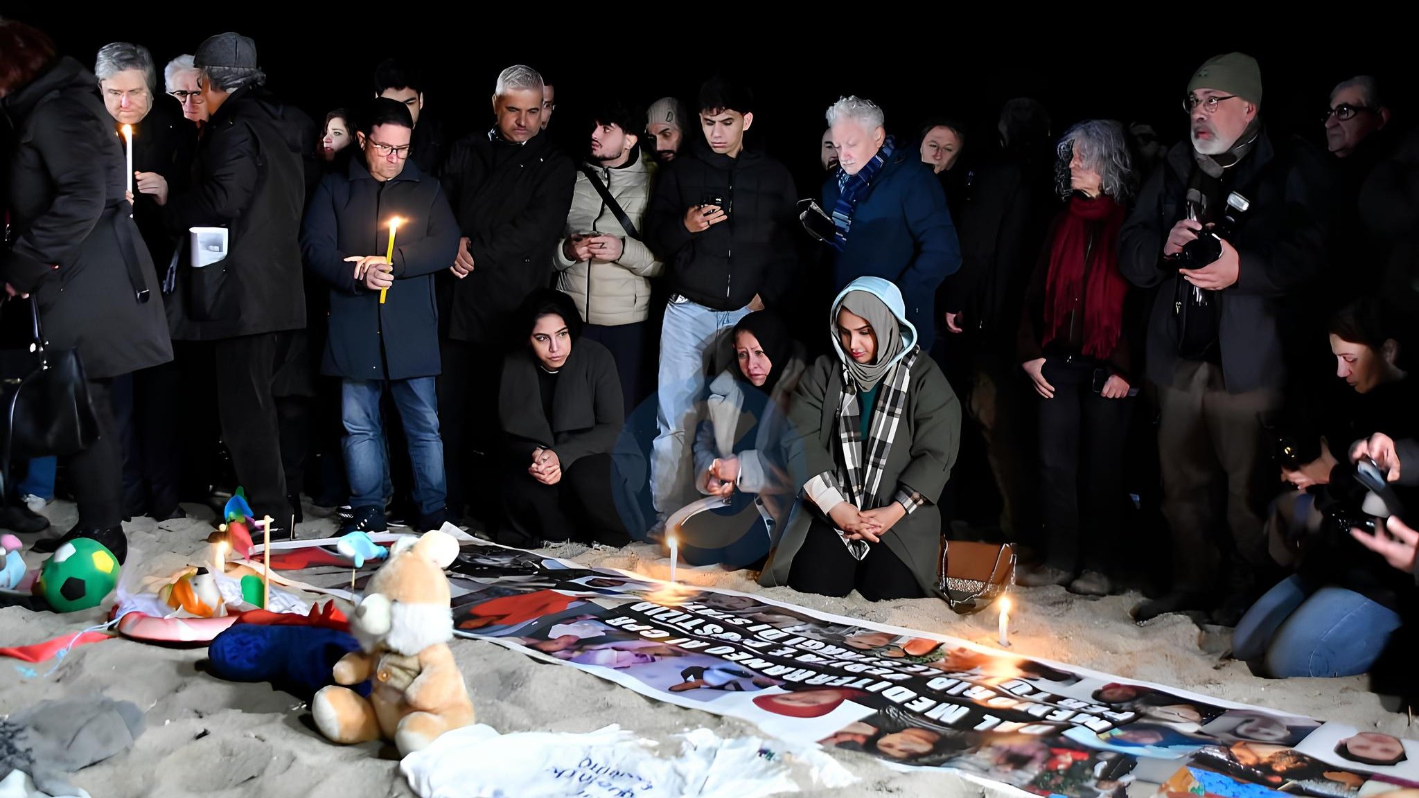 Un gruppo di persone in cerchio, con candele accese e striscioni, su una spiaggia al buio.