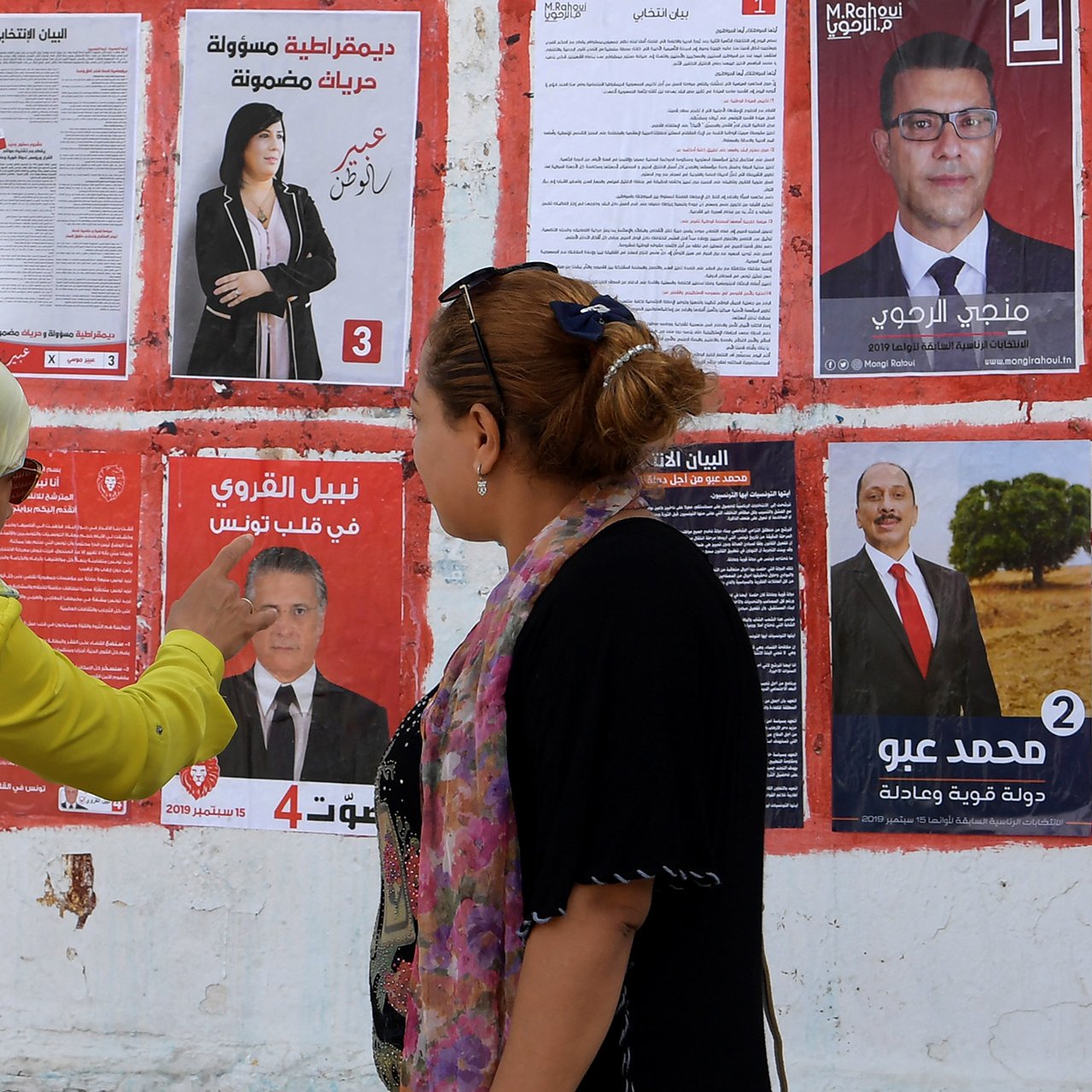 تظهر الصورة جدارًا مغطى بمجموعة من الملصقات الانتخابية. هناك سيدتان تقفان أمام الملصقات. إحداهما ترتدي عباءة ملونة وتظهر وهي تشير إلى أحد الملصقات. يتواجد ملصقات لمرشحين مختلفين، يحمل كل منها صورة واسم المرشح ورقمًا. تعكس الصورة أجواء الانتخابات والحماس المحيط بها، حيث تبدو السيدتان مهتمتين بمناقشة المعلومات المعروضة على الملصقات.