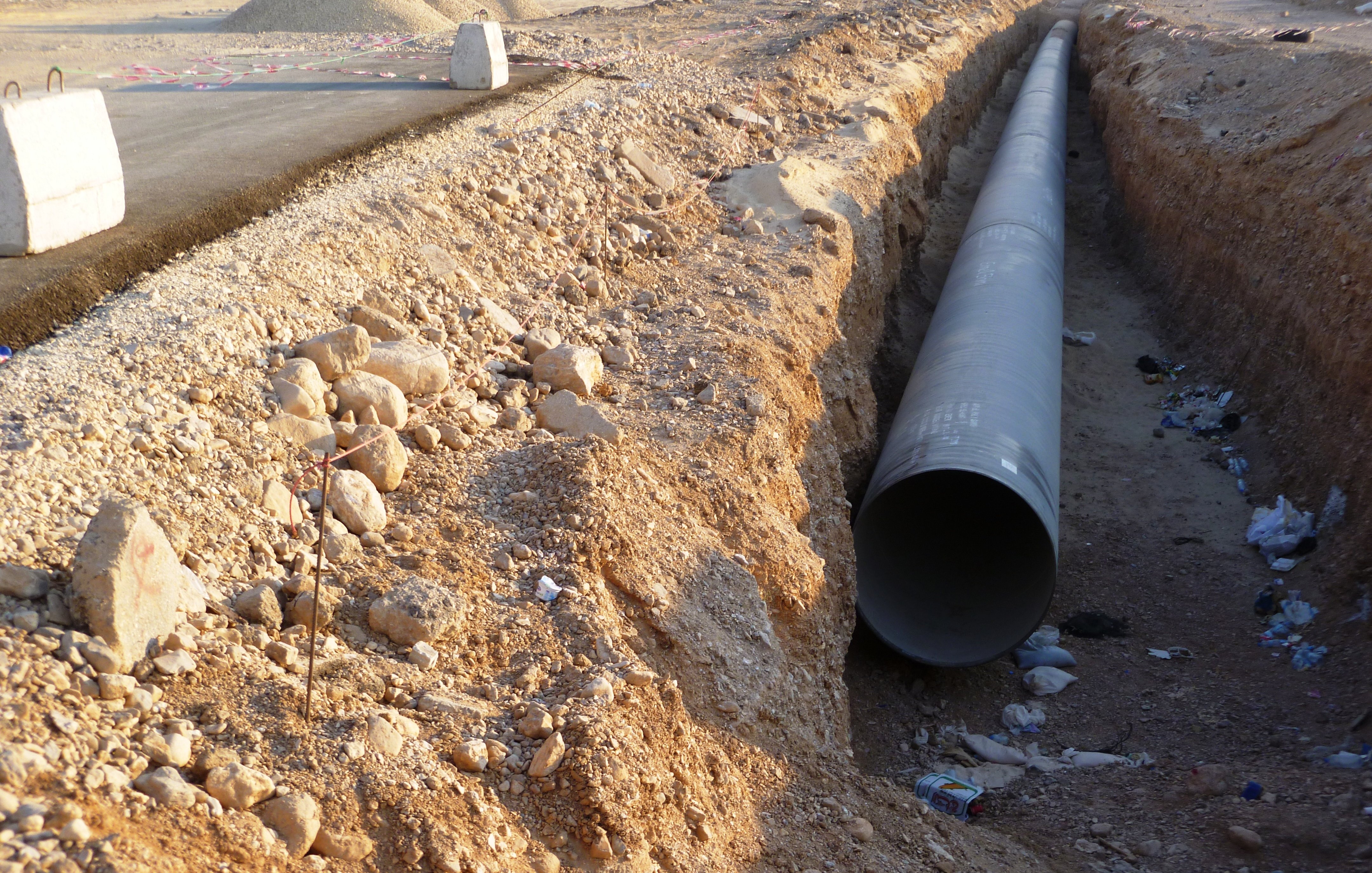 تظهر الصورة مشهدًا لموقع عمل حيث تم حفر خندق في أرض جافة. في وسط الخندق، يوجد أنبوب كبير على الأرض، ويبدو أنه يتم إعداده للتركيب. يحيط بالمنطقة بعض الحصى والصخور، مما يدل على أعمال البناء. في الخلفية، يمكن رؤية معدات بناء تظهر أنها تعمل في الموقع. كما يوجد بعض المخلفات والقمامة حول الخندق، مما يشير إلى تواجد نشاط بشري.