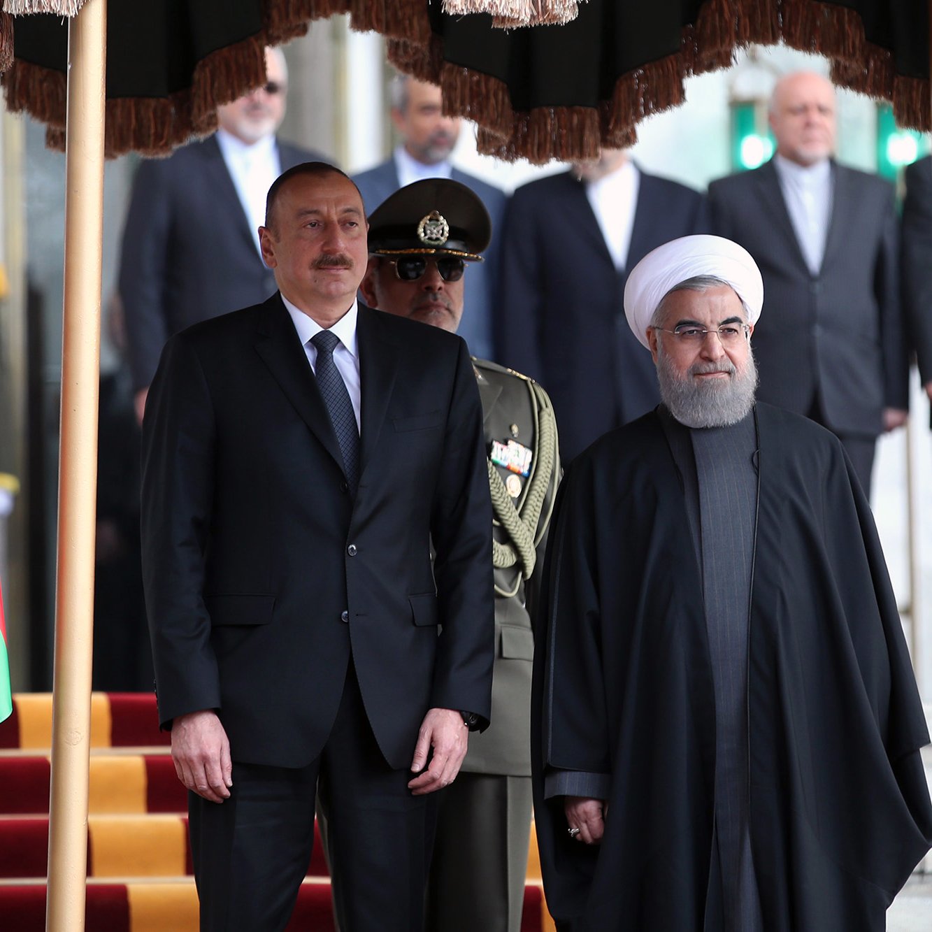 L'image montre deux hommes en costume, probablement des leaders politiques, posant ensemble devant un drapeau azéri. L'un d'eux porte un costume formel, tandis que l'autre est vêtu d'une tenue traditionnelle, avec une robe sombre. À l'arrière-plan, on aperçoit des soldats en uniforme et d'autres personnes, formant une sorte de cérémonie officielle. L'environnement semble être un bâtiment gouvernemental, avec des éléments décoratifs.