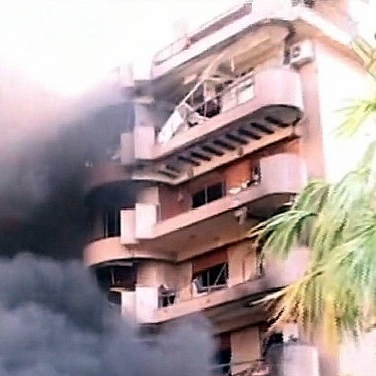 L'image montre un bâtiment en proie à un incendie. De la fumée noire s'échappe des fenêtres, tandis que l'extérieur du bâtiment semble endommagé. Des éléments de végétation, comme un palmier, sont visibles au premier plan, contrastant avec la scène chaotique et la destruction. Le ton général de l'image est sombre, mettant en évidence l'urgence et la gravité de la situation.