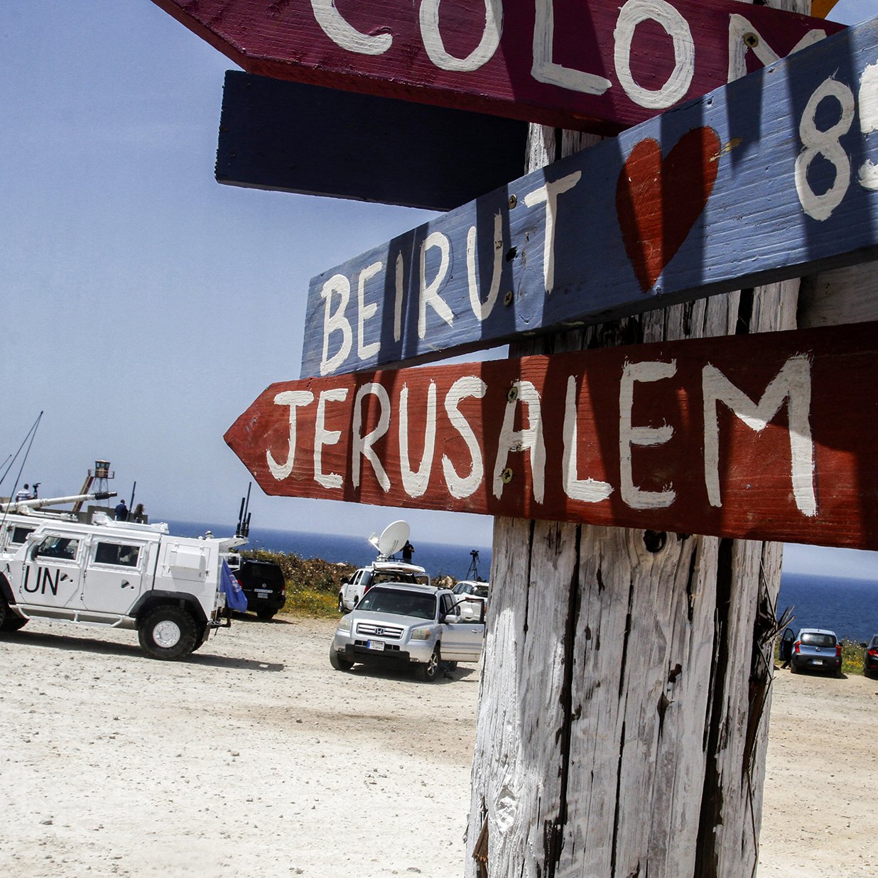 الصورة تظهر لافتة تشير إلى الاتجاهات والمسافات إلى عدة وجهات. هناك سهام ملونة تشير إلى مدن مختلفة مثل "بيروت" و"القدس"، مع ذكر المسافات بالكيلومترات. في الخلفية، يمكن رؤية مركبات، من ضمنها سيارة تحمل شعار الأمم المتحدة، مما يشير إلى وجود مهمة أو نشاط رسمي في المنطقة. المشهد يبدو هادئاً ويعكس طابعاً ساحلياً حيث يُرى البحر في الخلفية.