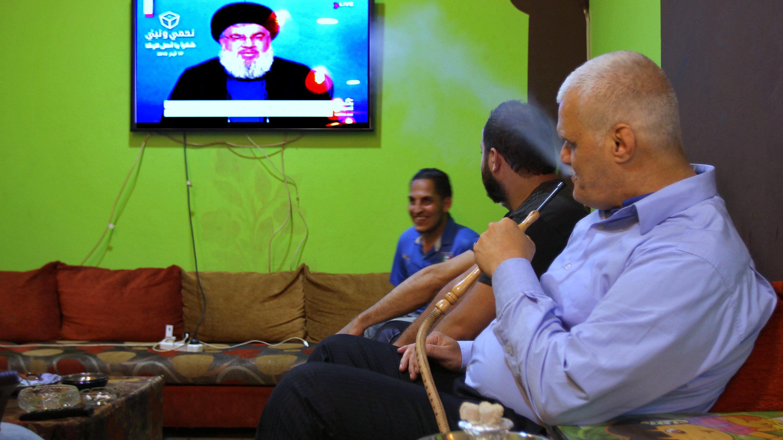 L'image montre un groupe d'hommes assis dans un salon avec des murs verts. Ils semblent regarder la télévision, où l'on peut voir un personnage à l'écran. L'un des hommes fume une pipe à eau, tandis que les autres sont engagés dans la conversation ou en observant l'écran. L'environnement est décontracté, avec des canapés confortables et des éléments de décoration simples.