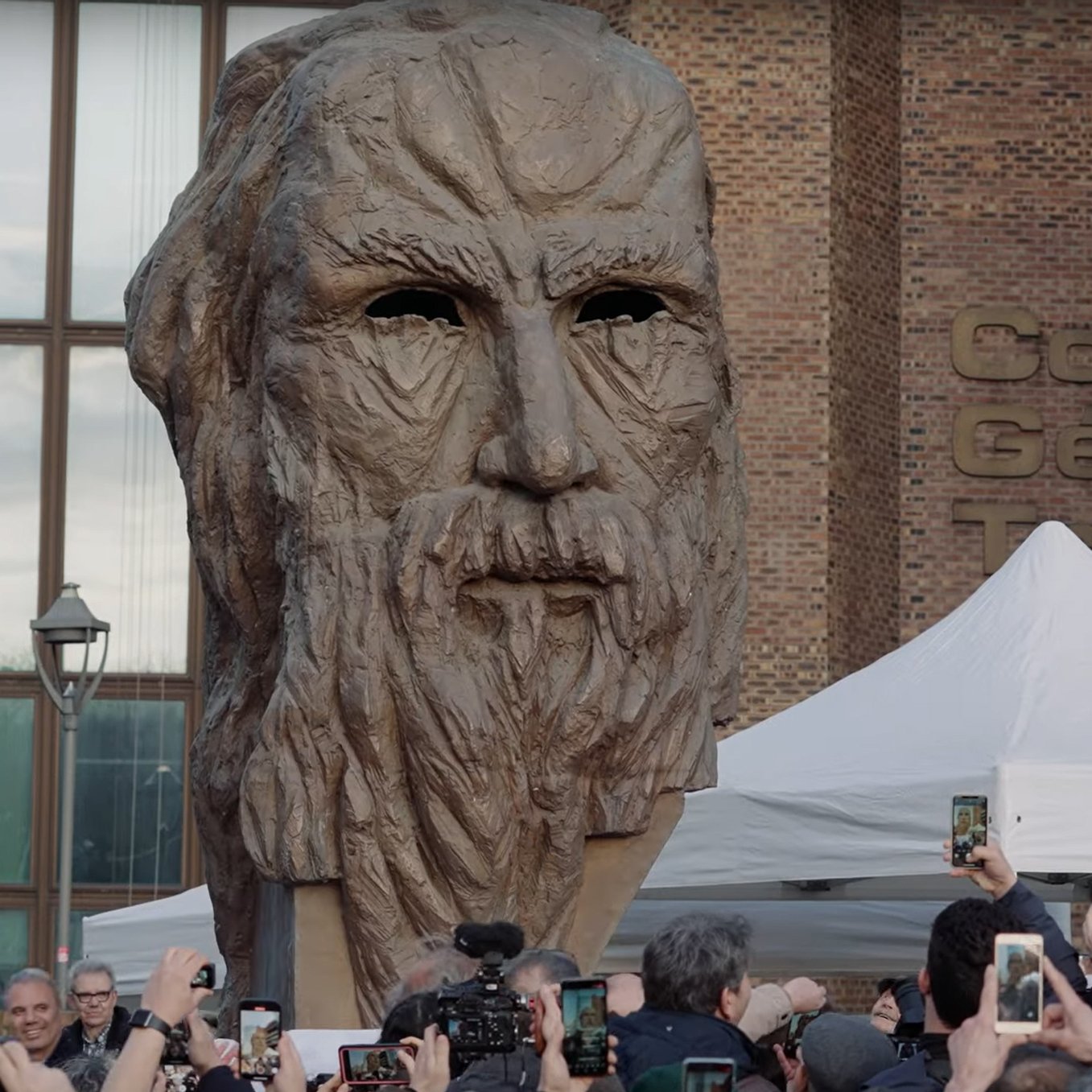L'image montre une grande sculpture représentant un visage ancien, probablement celui d'une figure historique ou mythologique, avec des traits marqués et une barbe. Elle est entourée d'une foule de personnes qui semblent prendre des photos avec leurs téléphones. Il y a aussi des tentes et un bâtiment visible en arrière-plan, ce qui indique qu'il s'agit probablement d'un événement public ou d'une exposition. L'atmosphère semble être festive et attentive, centrée sur l'œuvre sculpturale.