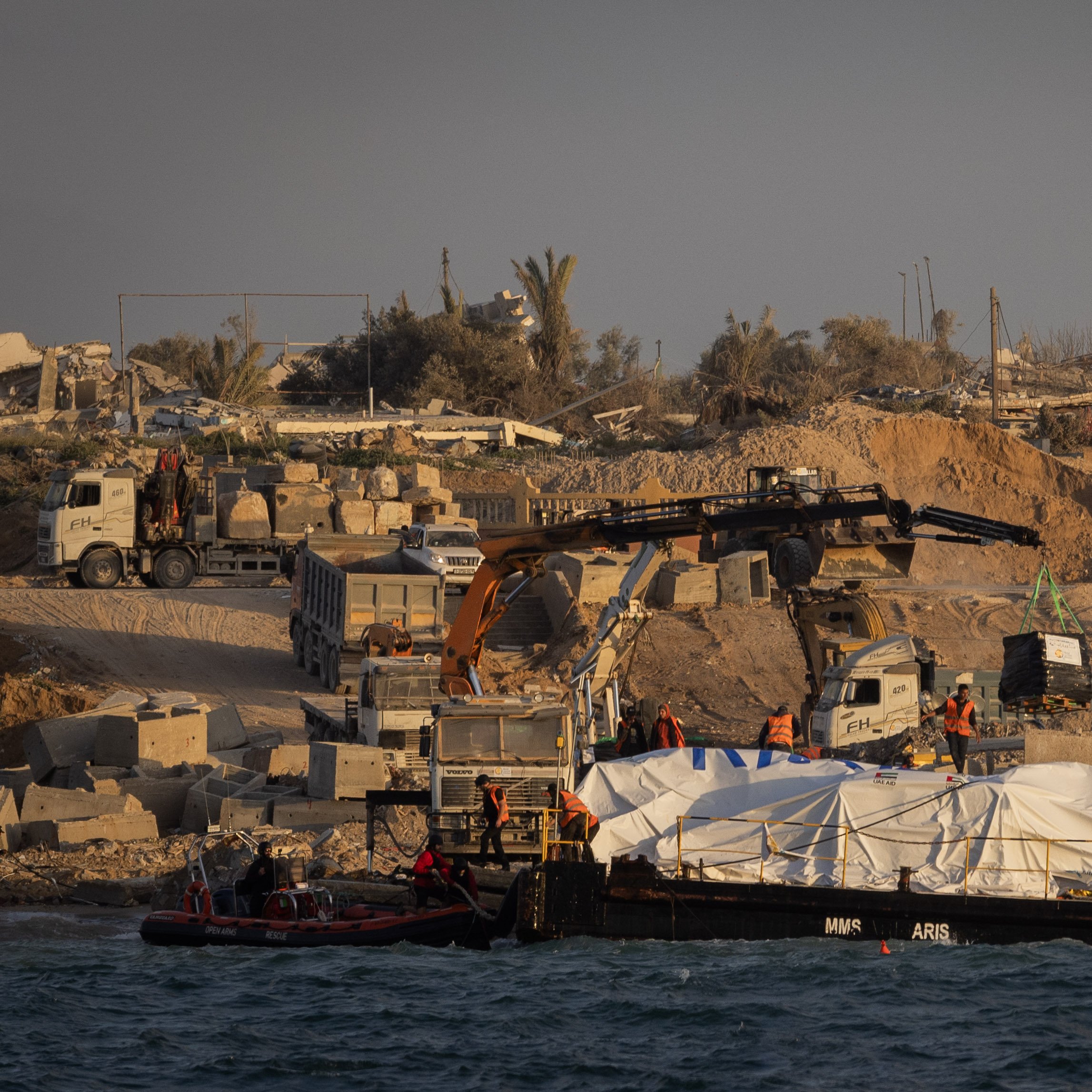 در این تصویر، یک صحنه ساختمانی در کنار دریا دیده می‌شود. در ساحل، تعدادی ماشین سنگین و جرثقیل در حال کار هستند. یک قایق روی آب قرار دارد که زیر آن یک پوشش سفید رنگ قرار دارد. پس‌زمینه شامل ساختمان‌های نیمه‌کار و تخریب شده و چند درخت نخل است. نور خورشید به صورت گرم بر روی این صحنه تابیده و حال و هوای آن را به تصویر کشیده است.